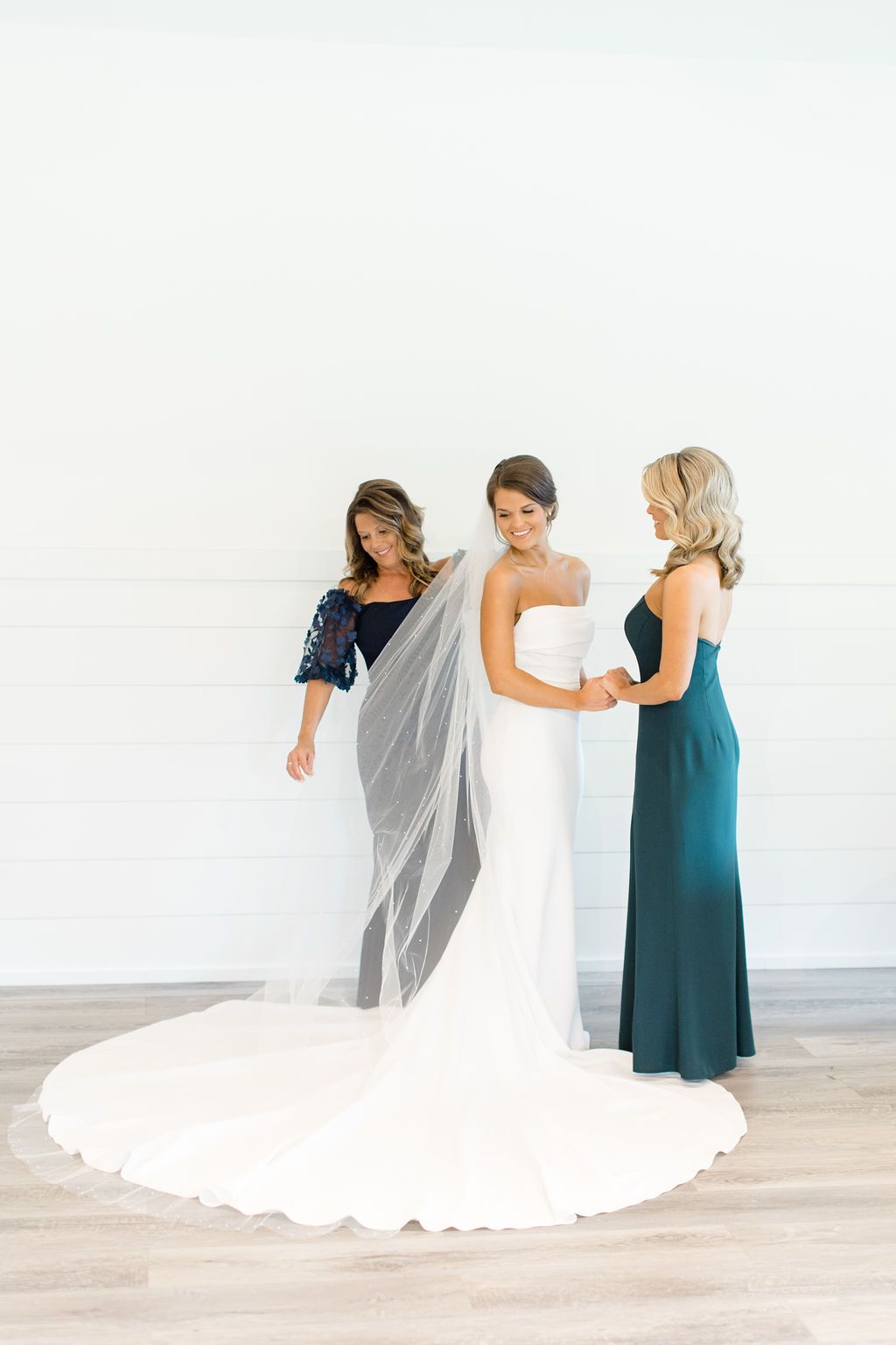 Three women in formal dresses stand together in a bright room, one in a white gown with a long train.