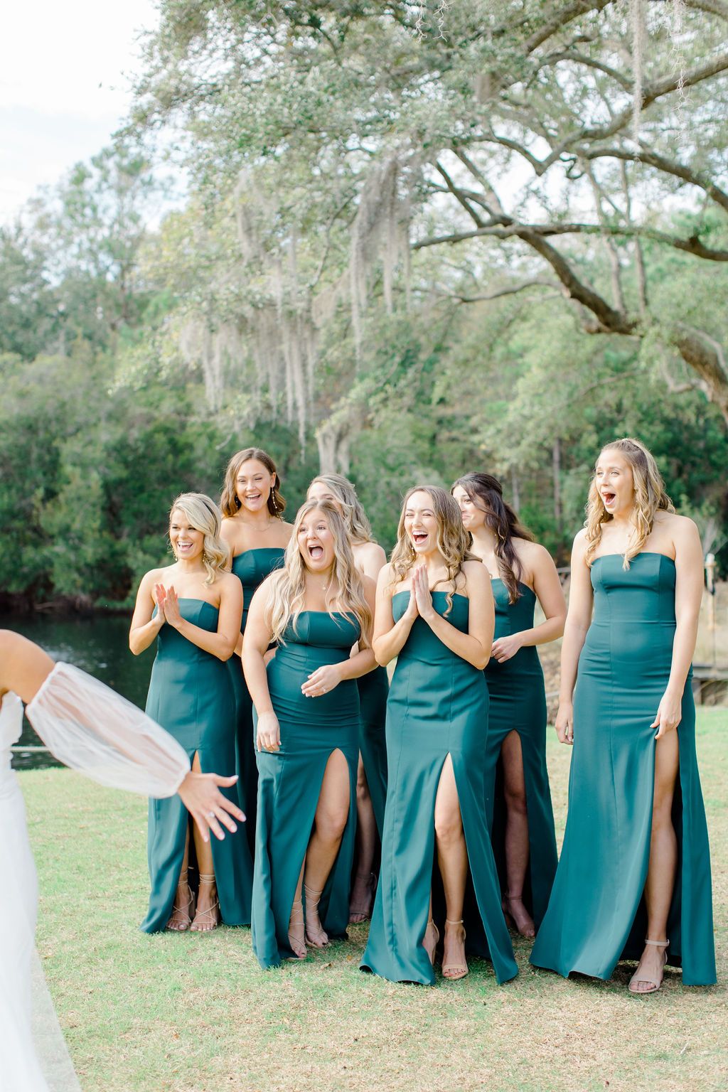 Group of women in teal dresses posing outdoors on grass beneath large trees.