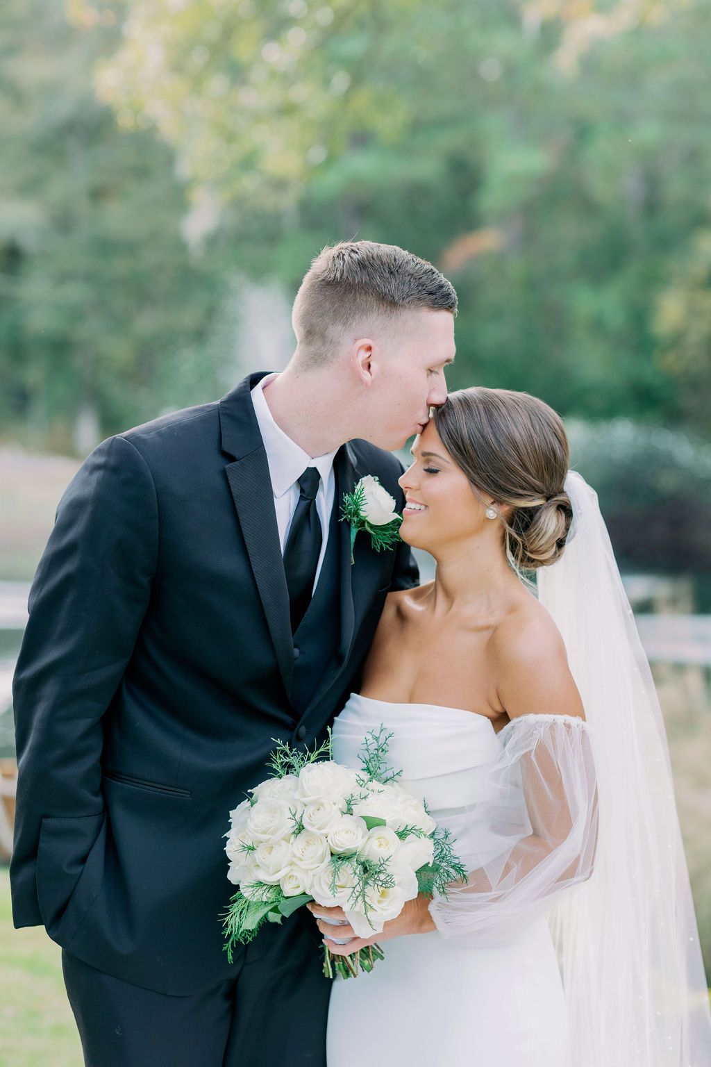 Bride and groom sharing a kiss outdoors, bride holding a white bouquet in a white gown and veil.