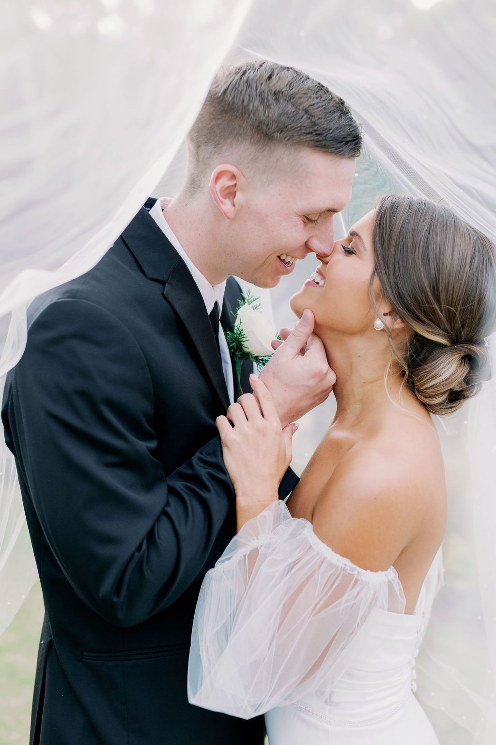 Bride and groom embracing under flowing white fabric, smiling and about to kiss at their wedding