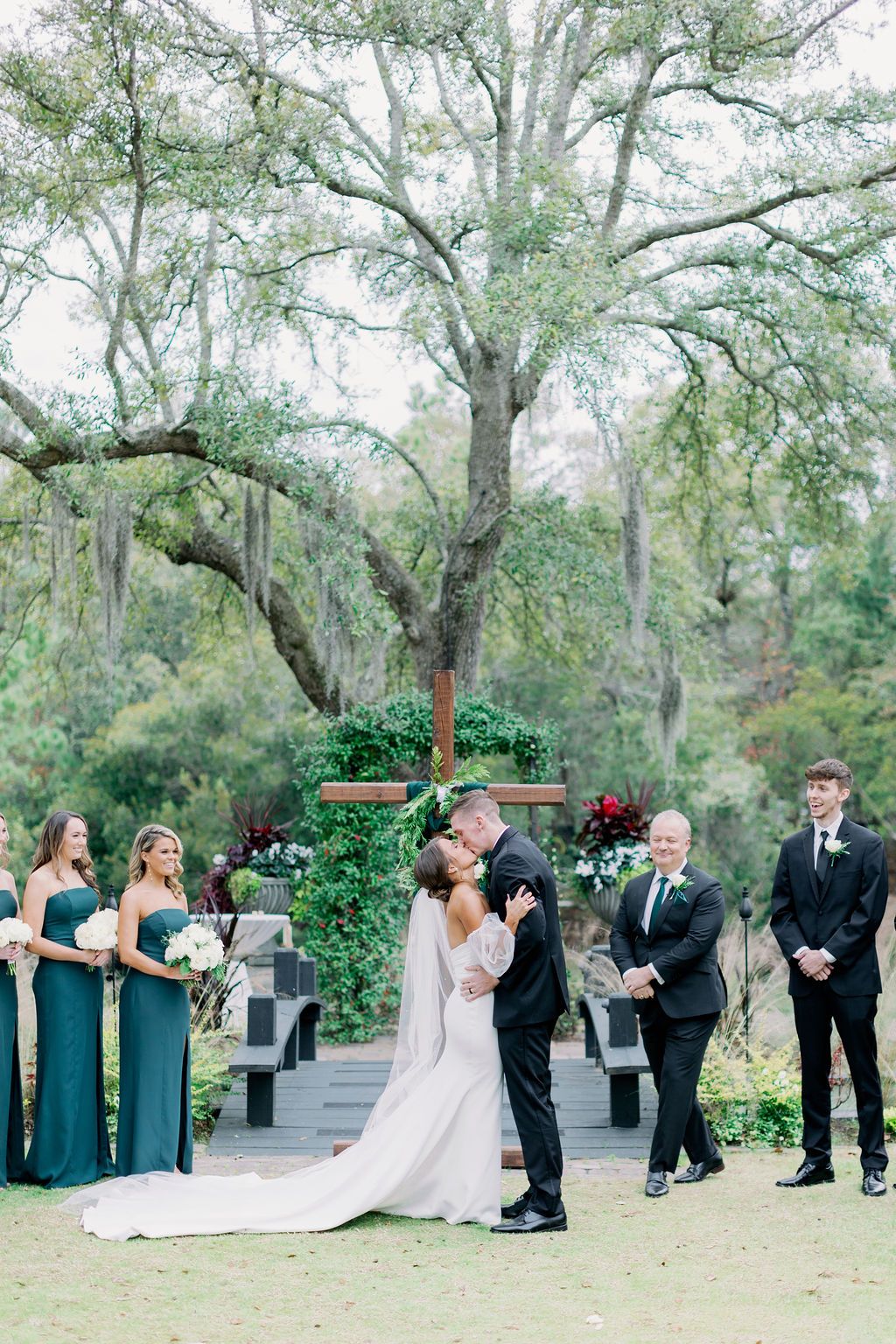Bride and groom exchanging vows outdoors beneath tall trees, with wedding party nearby