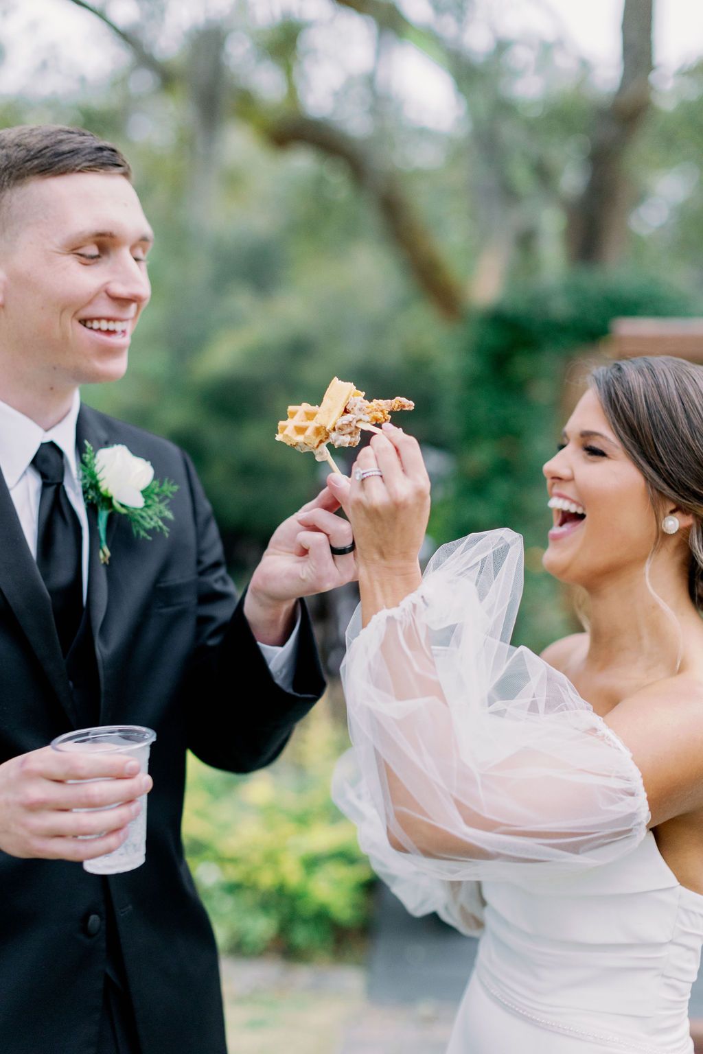 Bride and groom laugh and toast with drinks during an outdoor wedding celebration