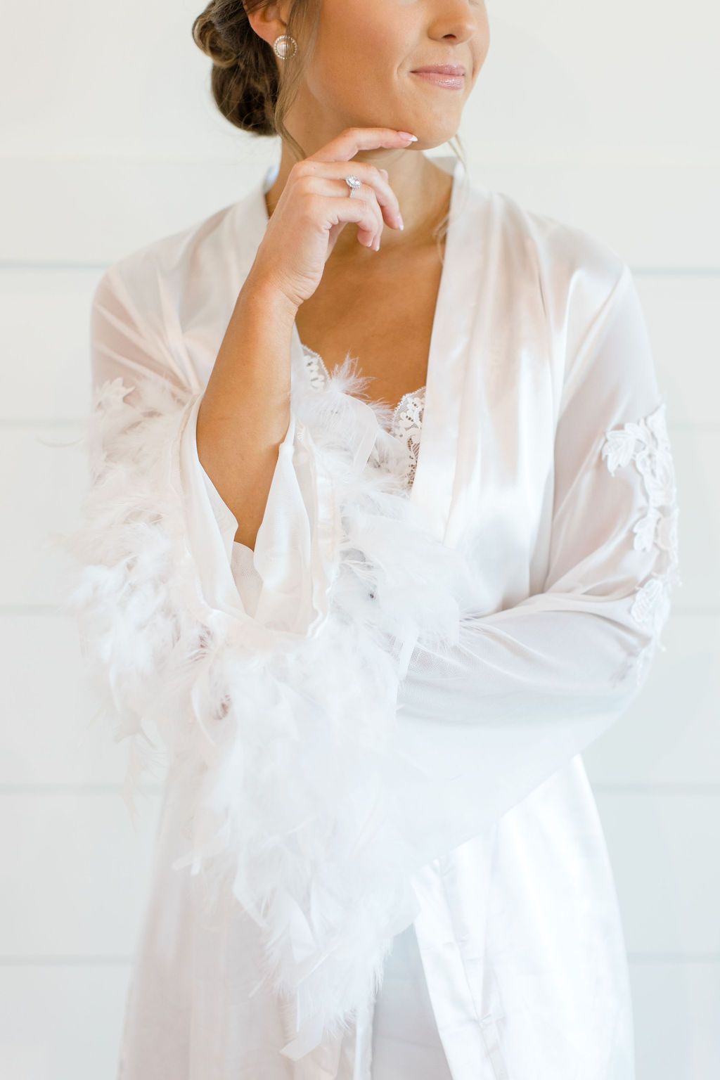 Woman in a white robe holding glasses near a lace-trimmed neckline, standing by a bright wall.