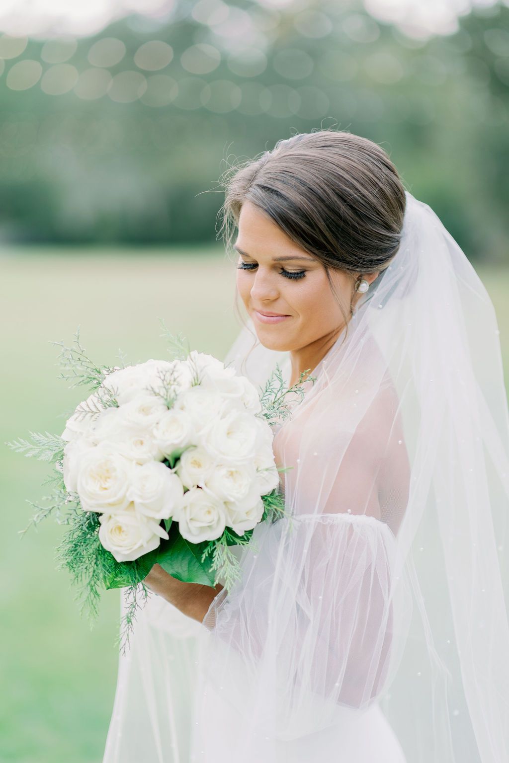 Bride holding a white bouquet in a white veil, standing outdoors with a soft green background