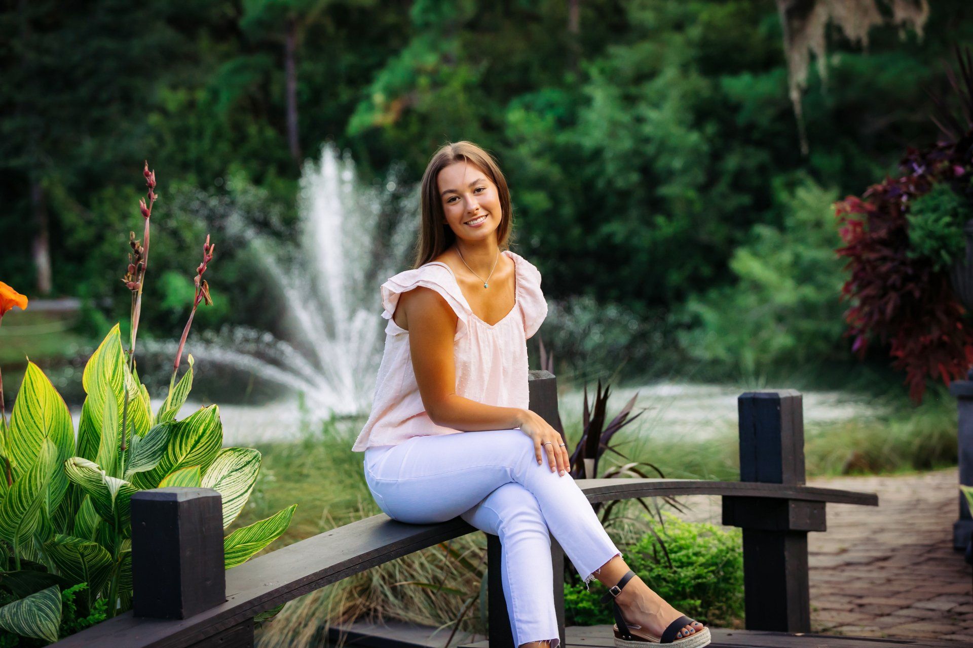 Woman in white sitting on a bench in a garden with a fountain and lush greenery in the background
