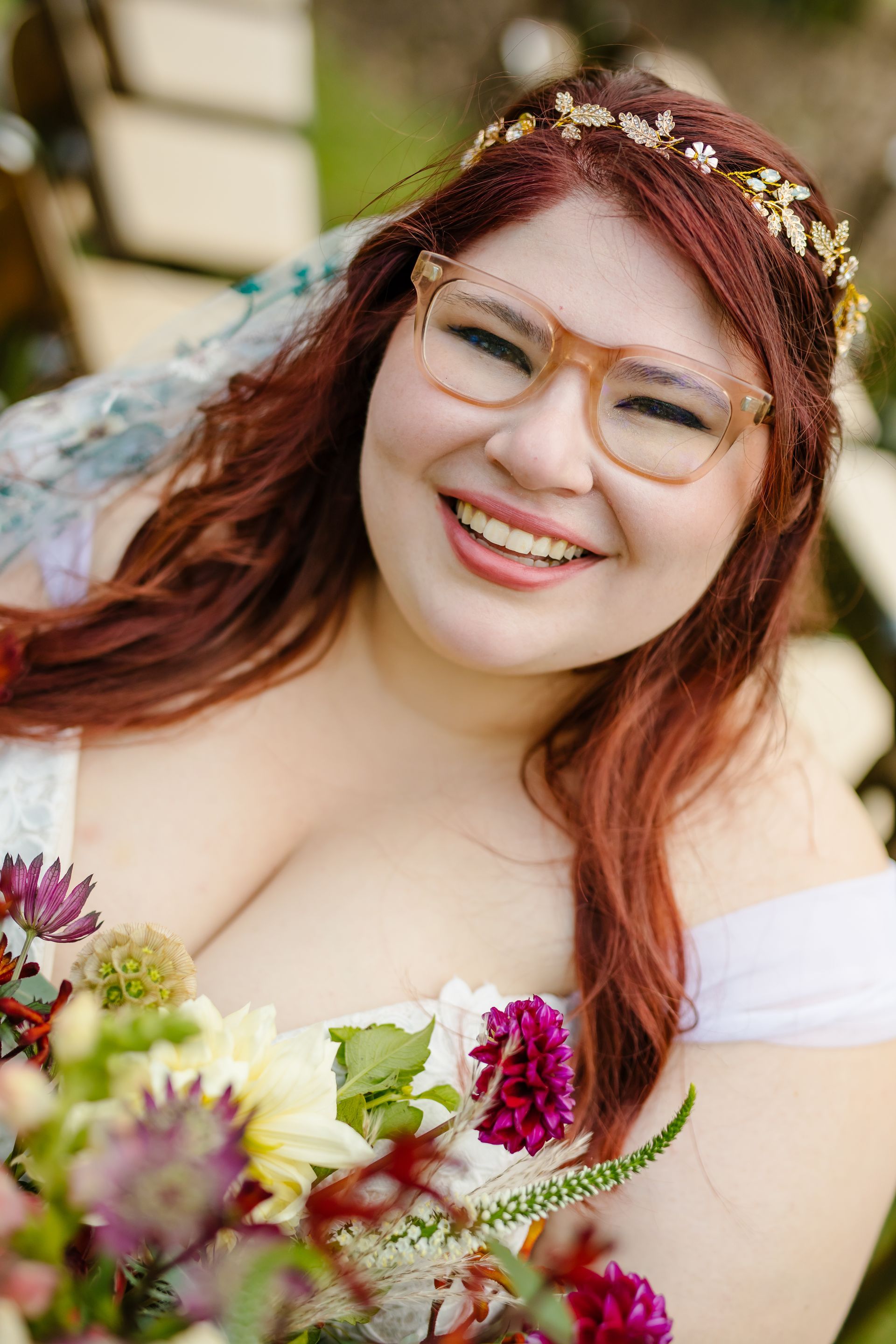 Smiling person with red hair and glasses, wearing a floral headband and holding a bouquet outdoors