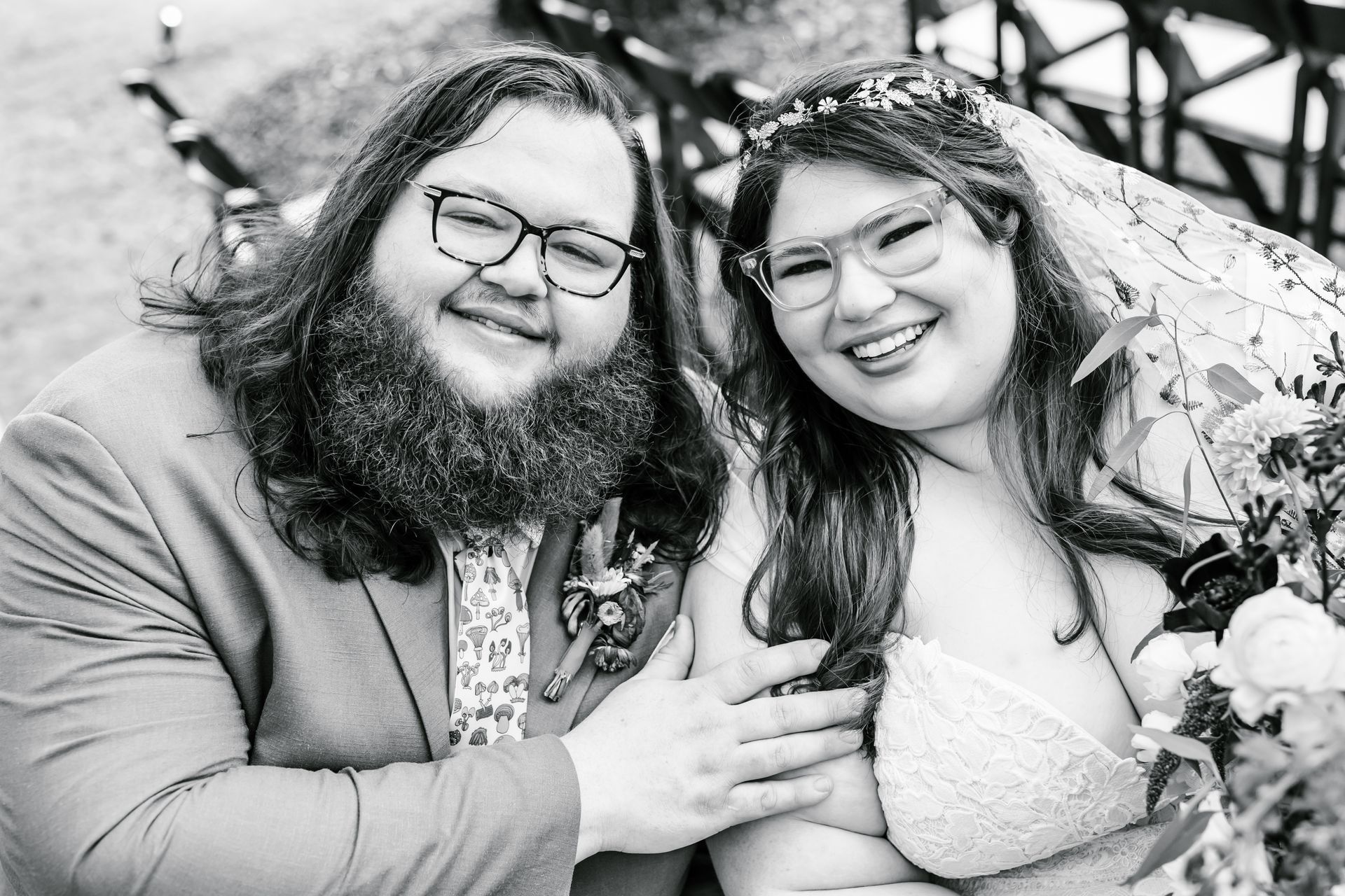Smiling couple posing outdoors in formal attire, holding flowers and wearing glasses.
