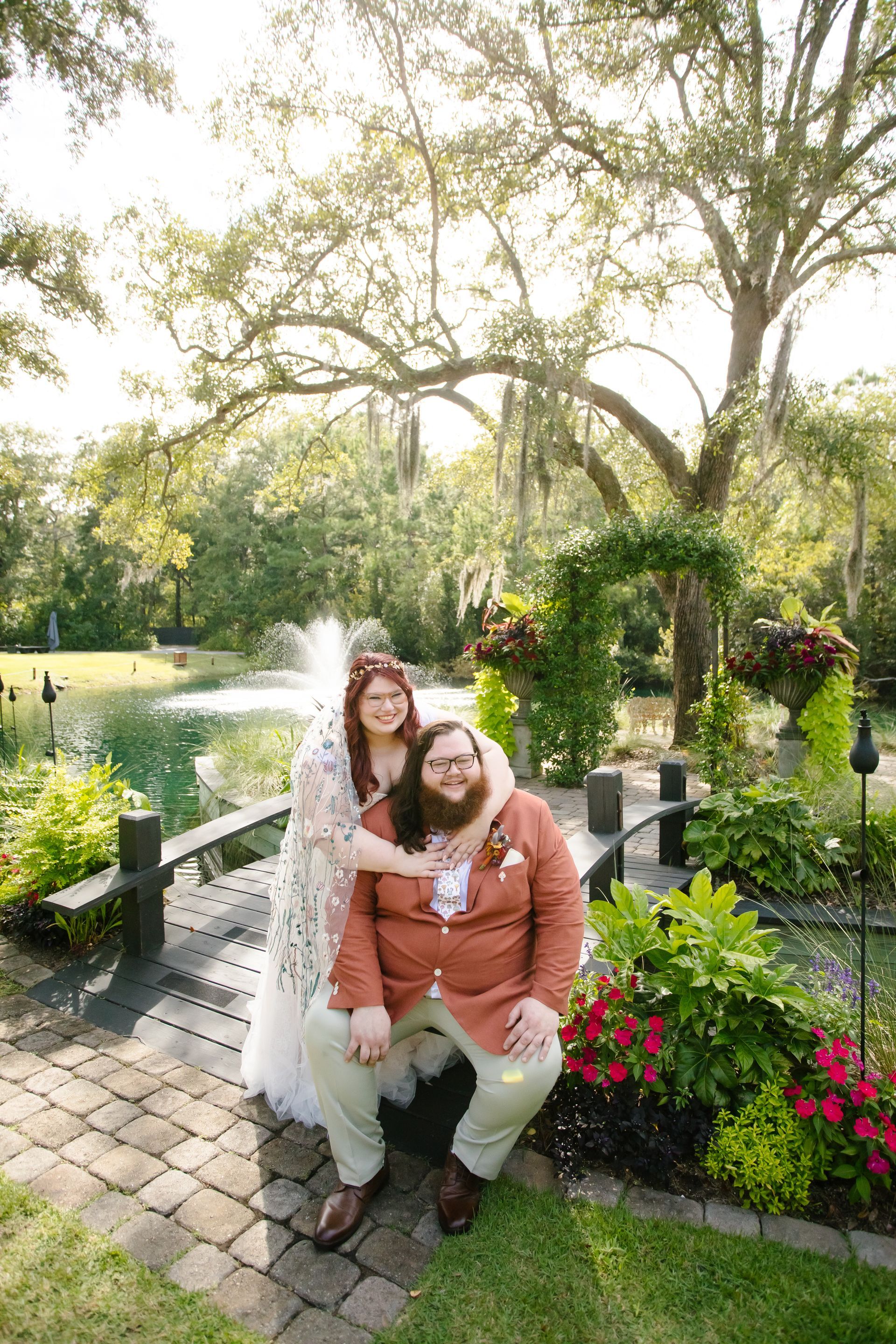 Two people posing on a bench beside a pond, surrounded by flowers and trees in a sunny garden.
