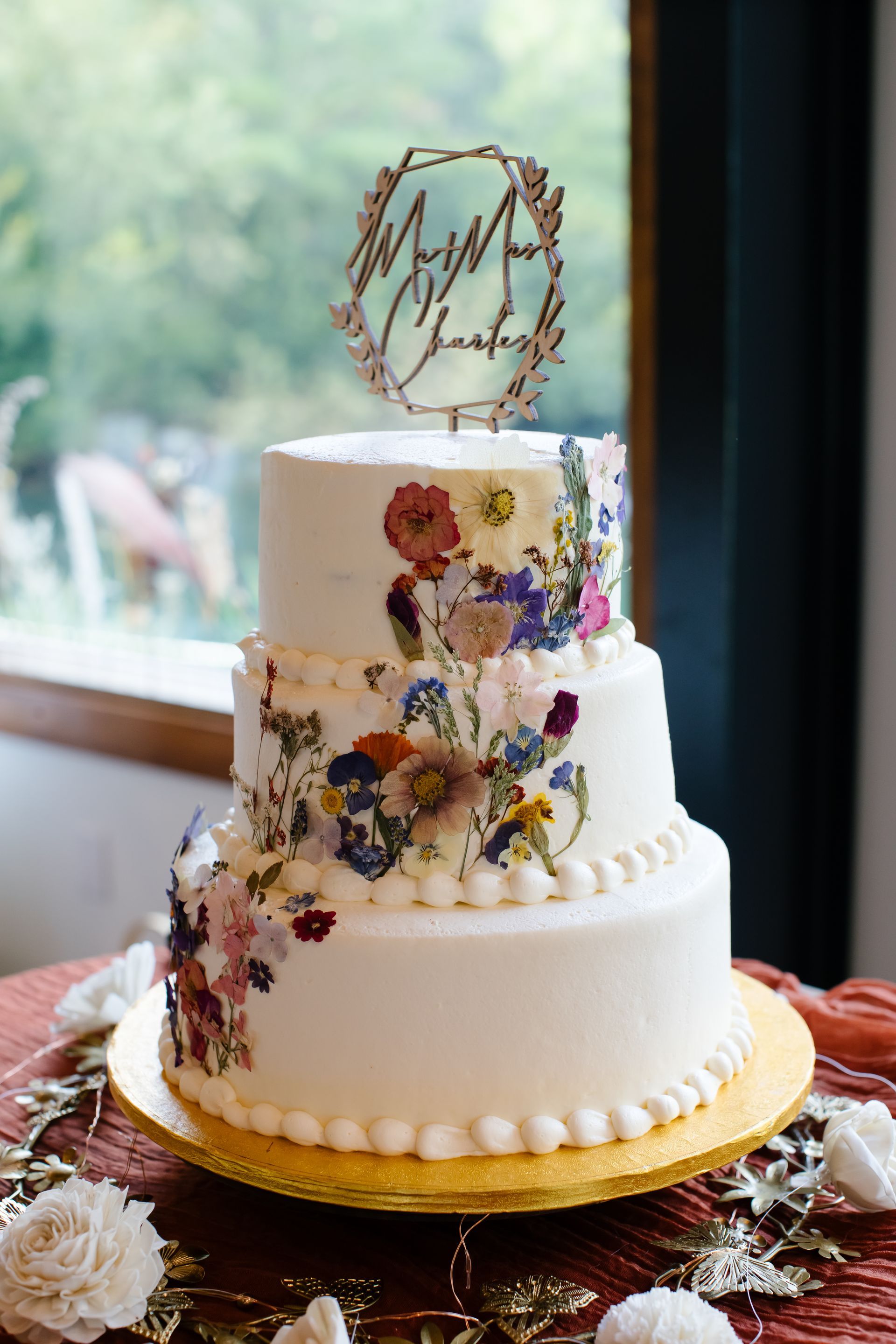 Three-tier floral wedding cake with white frosting and gold topper on a table by a window