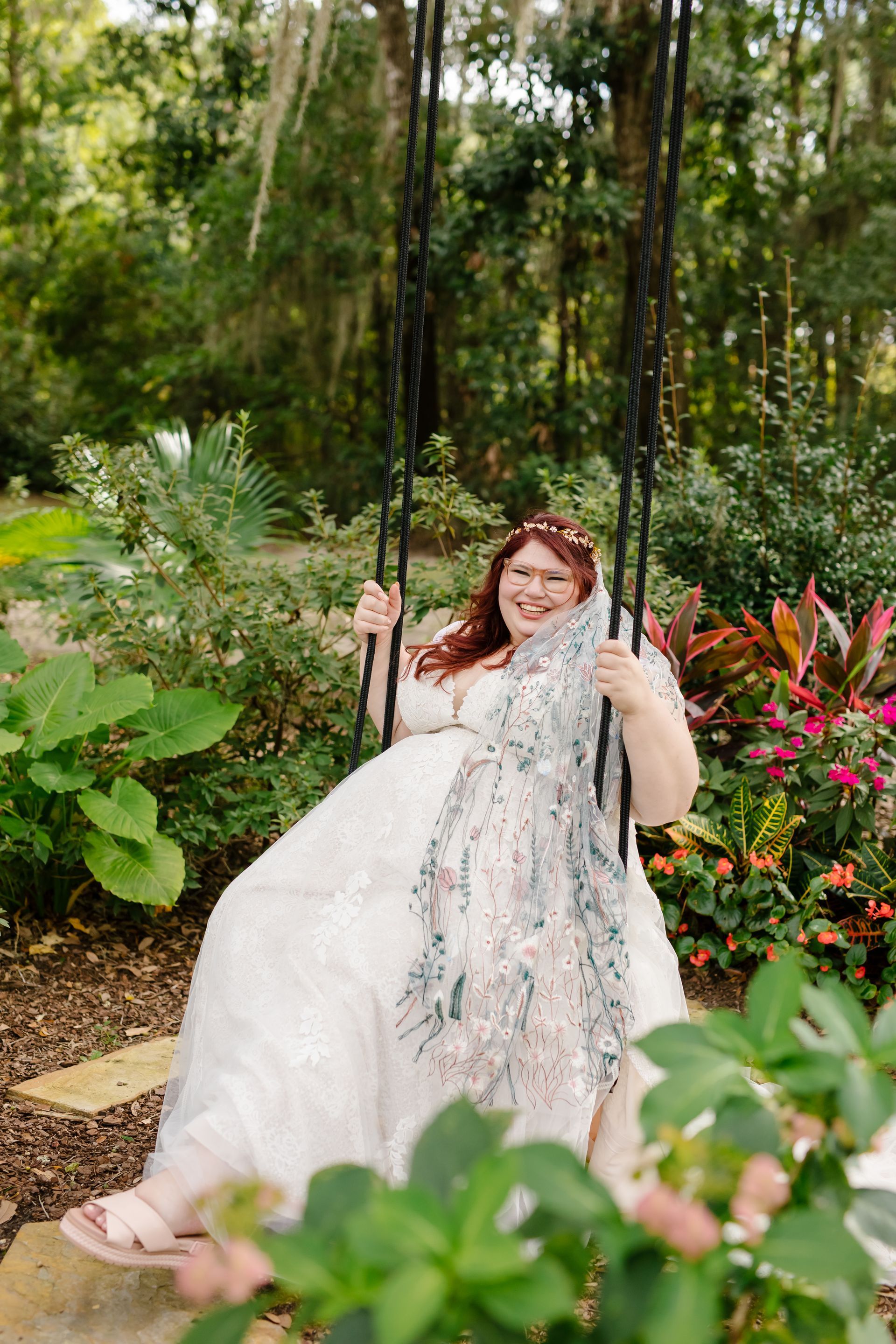 Person in a white dress smiling on a swing in a lush garden with colorful flowers.