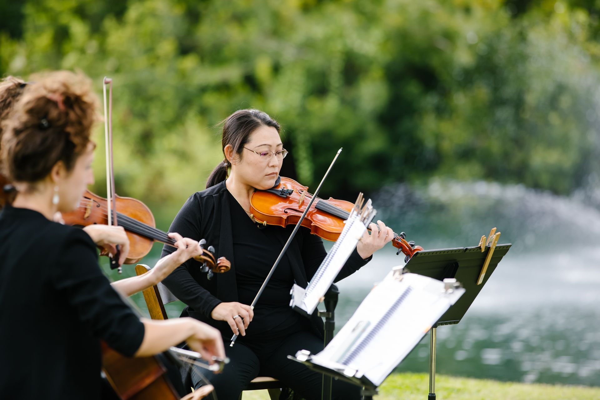 Musicians playing violins outdoors with sheet music stands by a lakeside