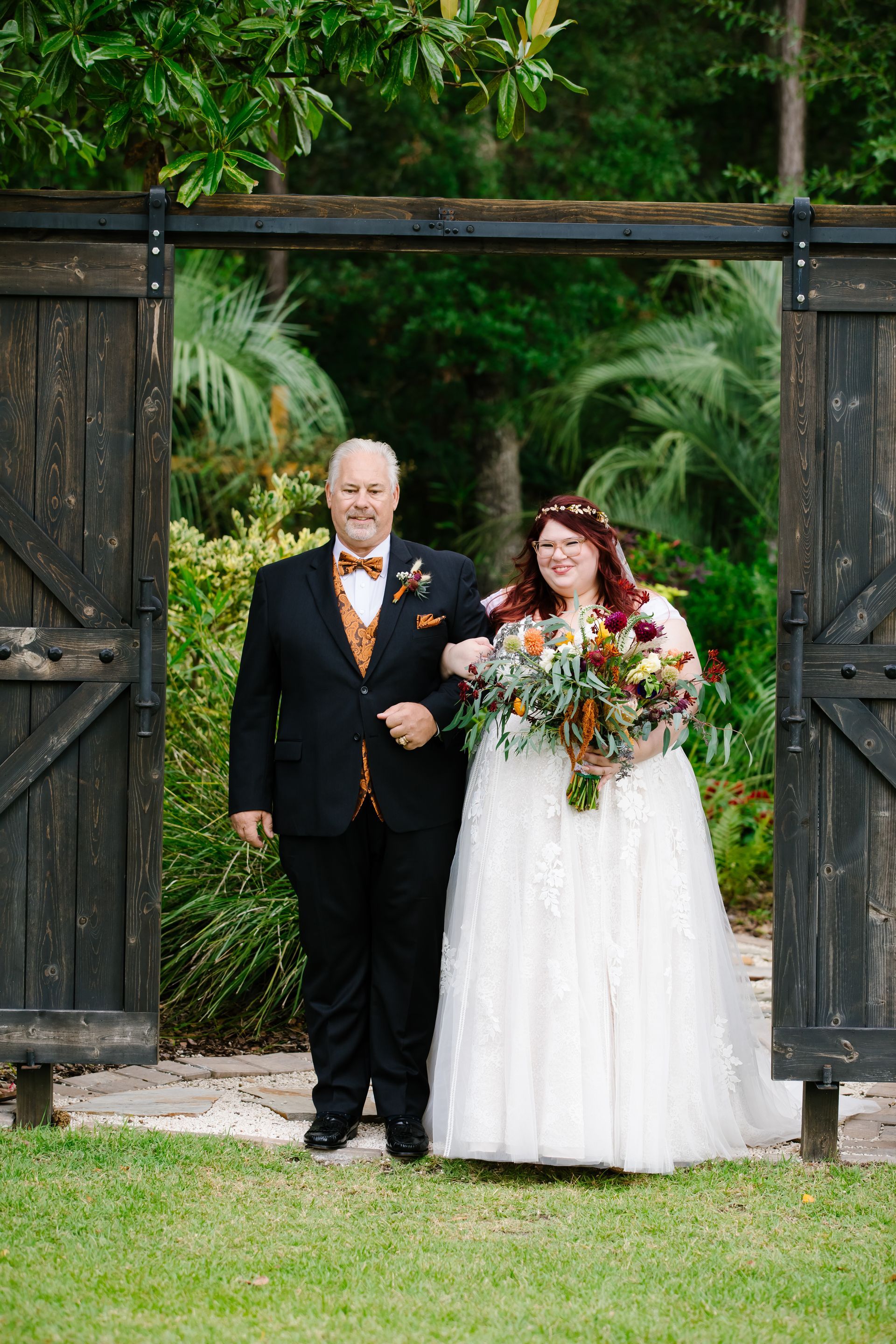 Bride and groom standing under a wooden archway in a garden, holding a bouquet with lush greenery behind them.