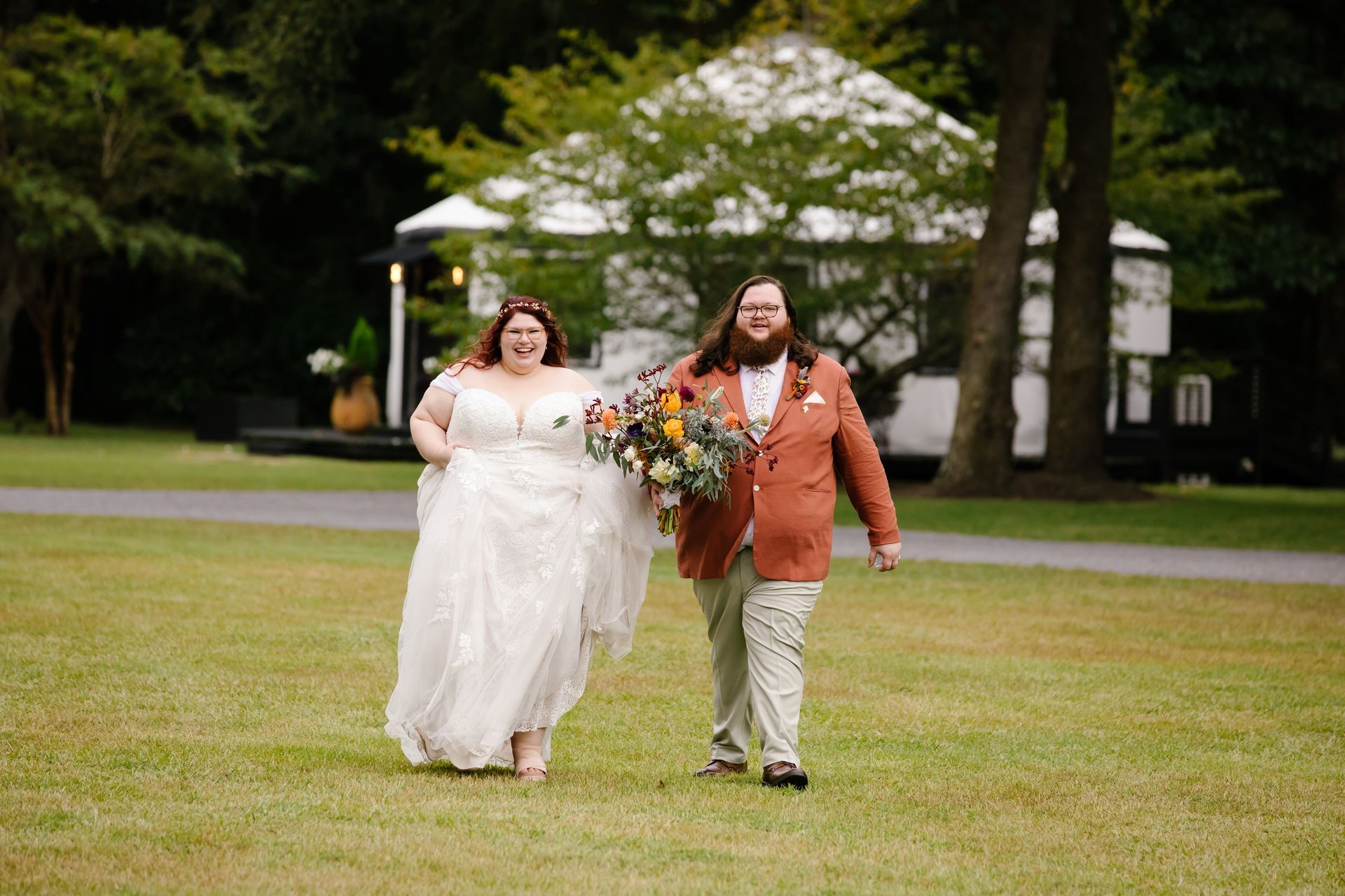 Two people in wedding attire walking across a grassy park, holding a bouquet.