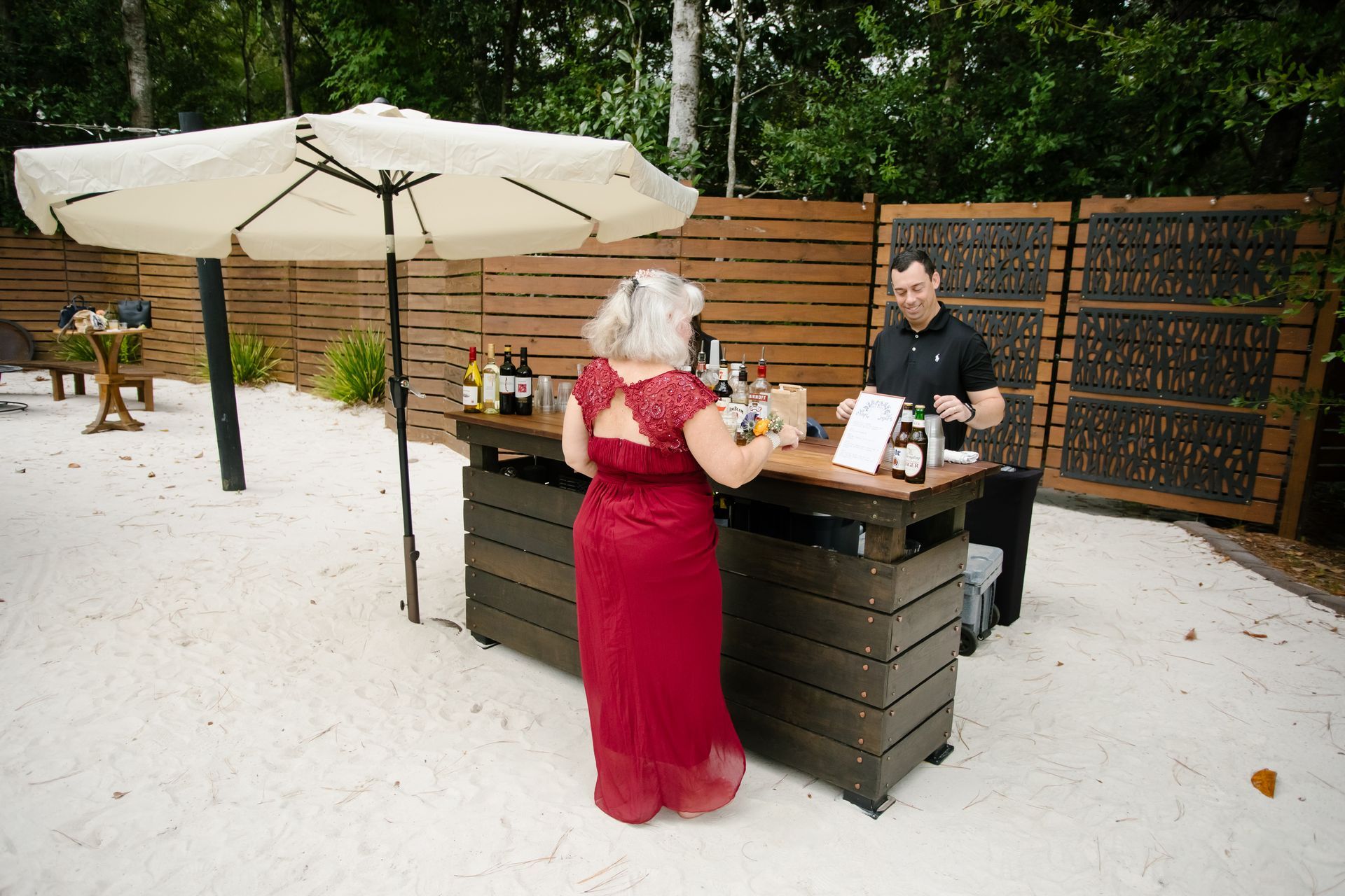 Woman in red dress at outdoor bar under large umbrella, speaking with bartender in a garden setting