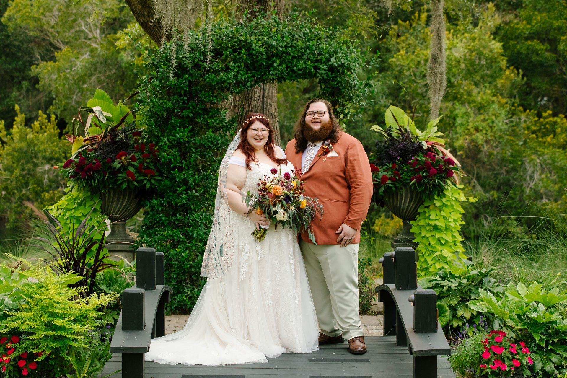 Couple in wedding attire posing on a garden bridge with a bouquet and lush greenery behind them