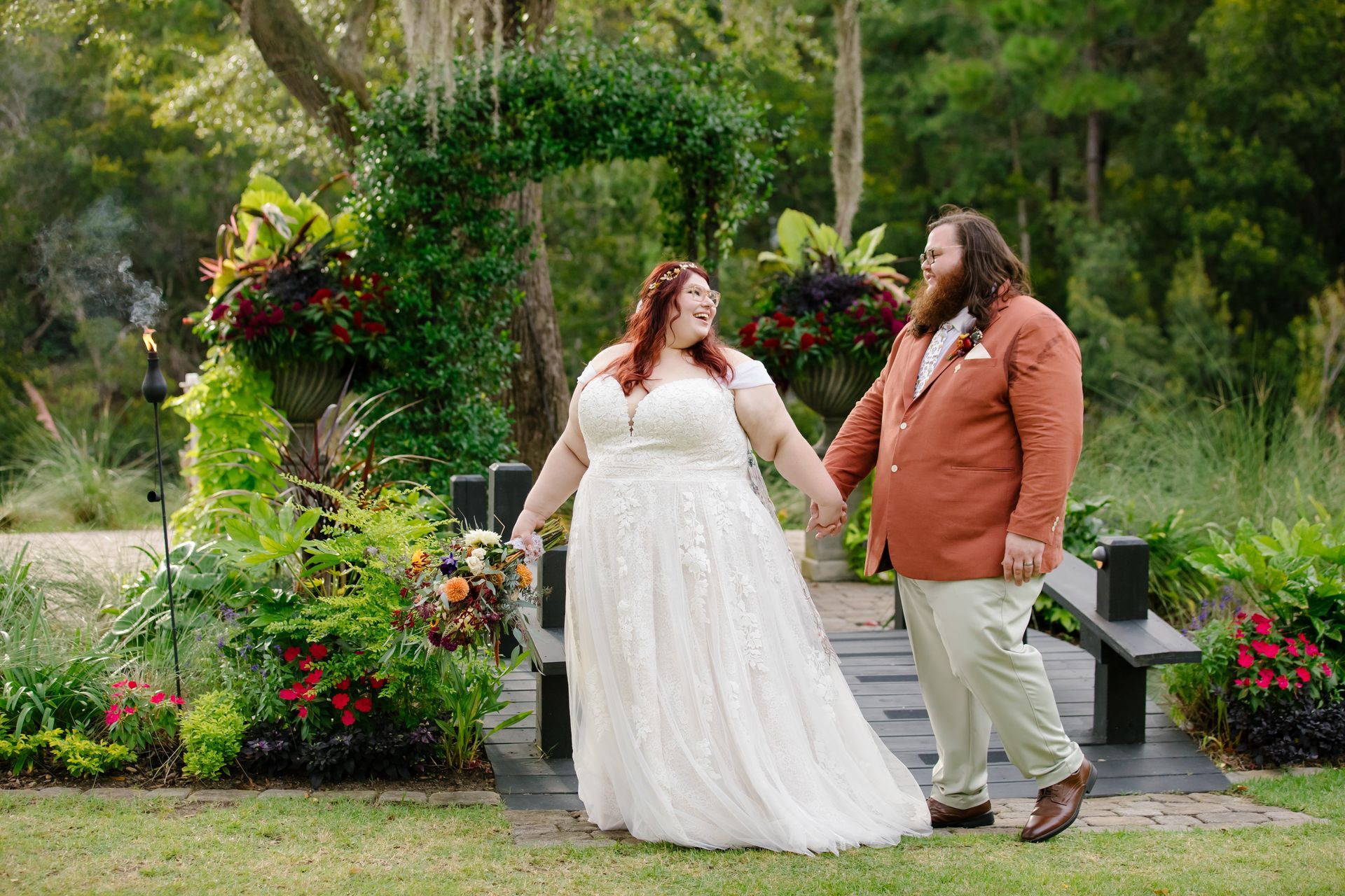 Couple in formal attire holding hands in a lush garden, smiling beside colorful flowers and steps.