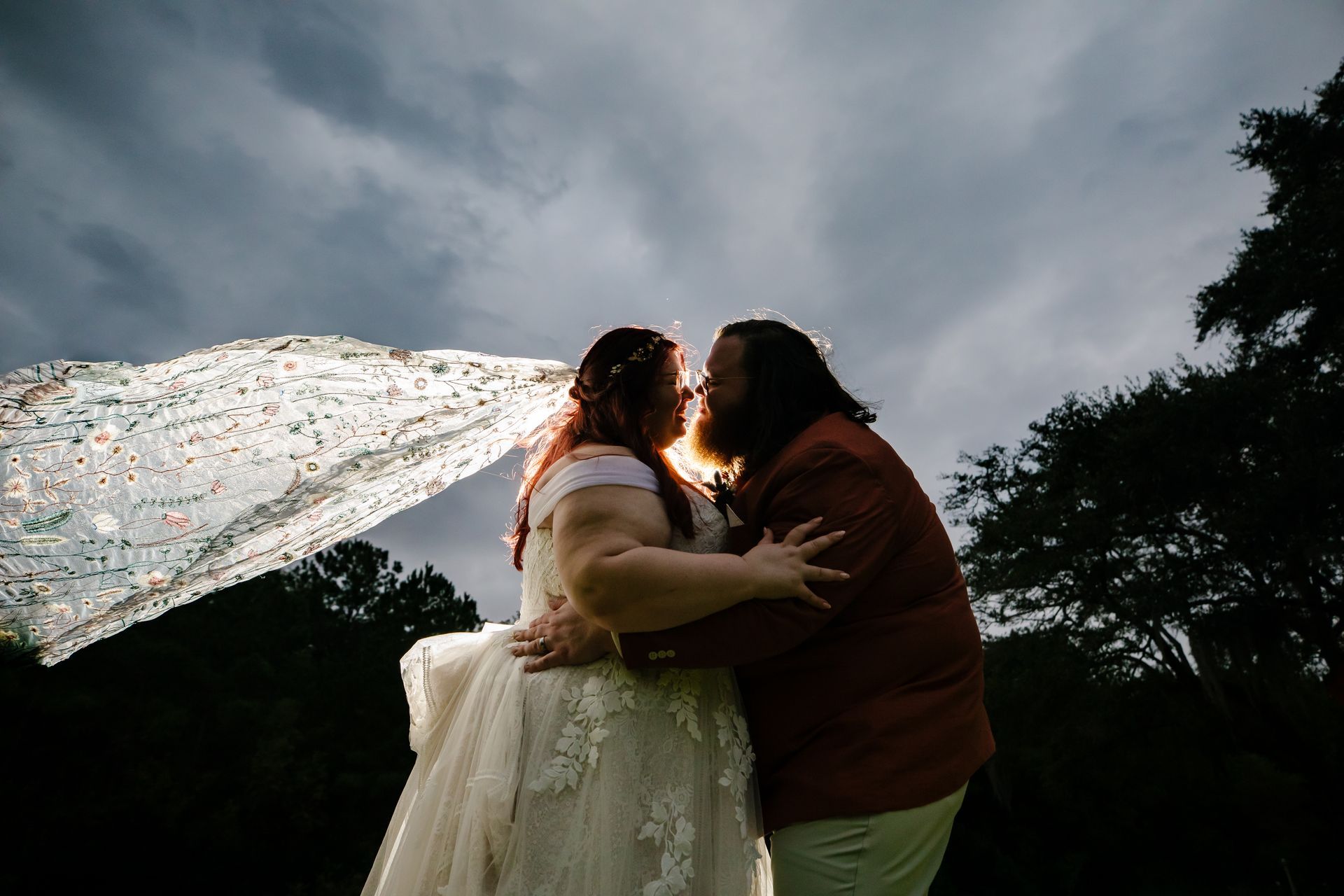 Two people embracing outdoors beneath a cloudy sky, with a flowing veil behind them.
