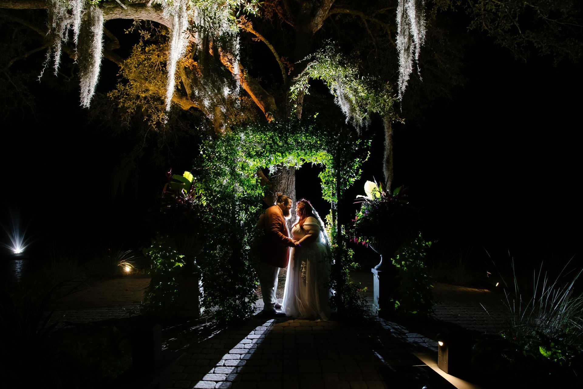 Couple standing under a leafy archway at night, lit by warm string lights in a garden path