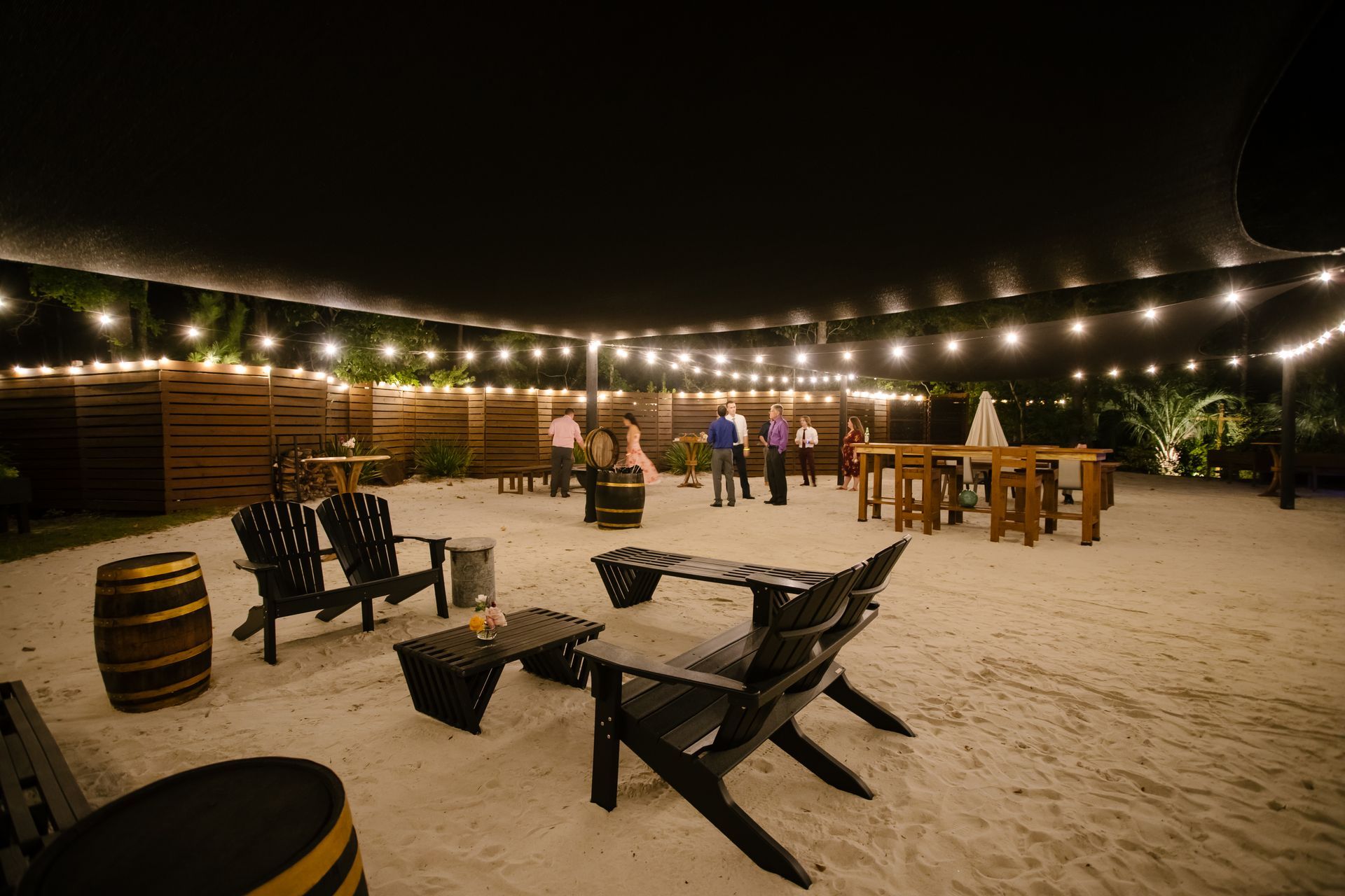 Outdoor nighttime beach venue with string lights, picnic tables, and a few people gathered on the sand