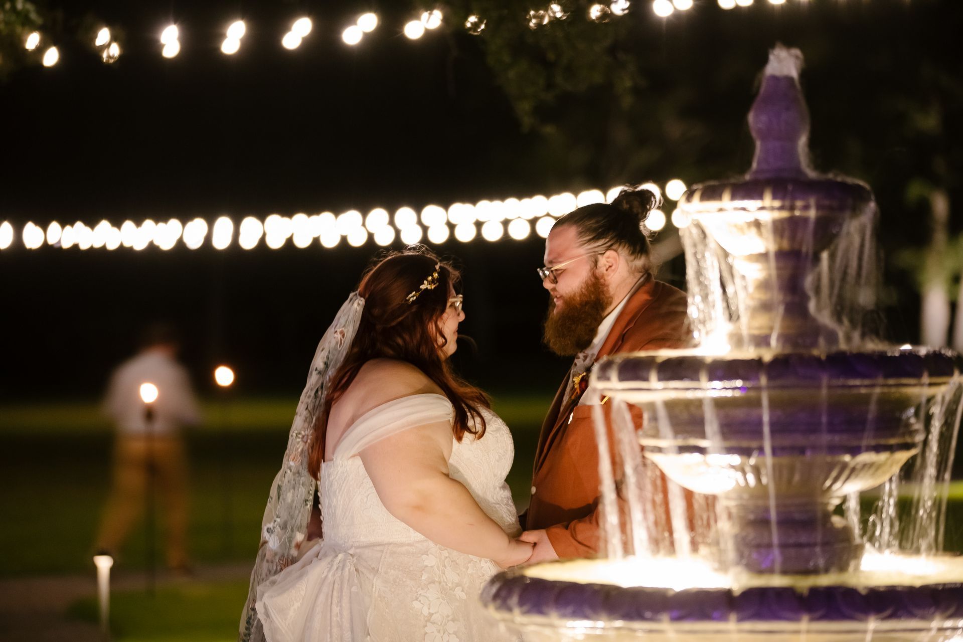 Couple standing beside a lit fountain at dusk, with string lights glowing in the background