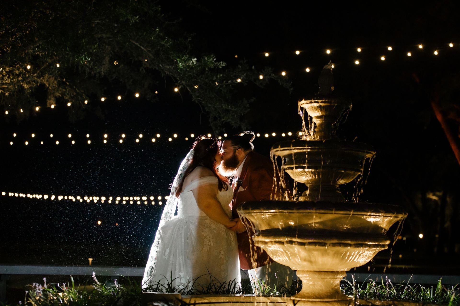 Couple kissing beside a lit fountain at night, with string lights in the background