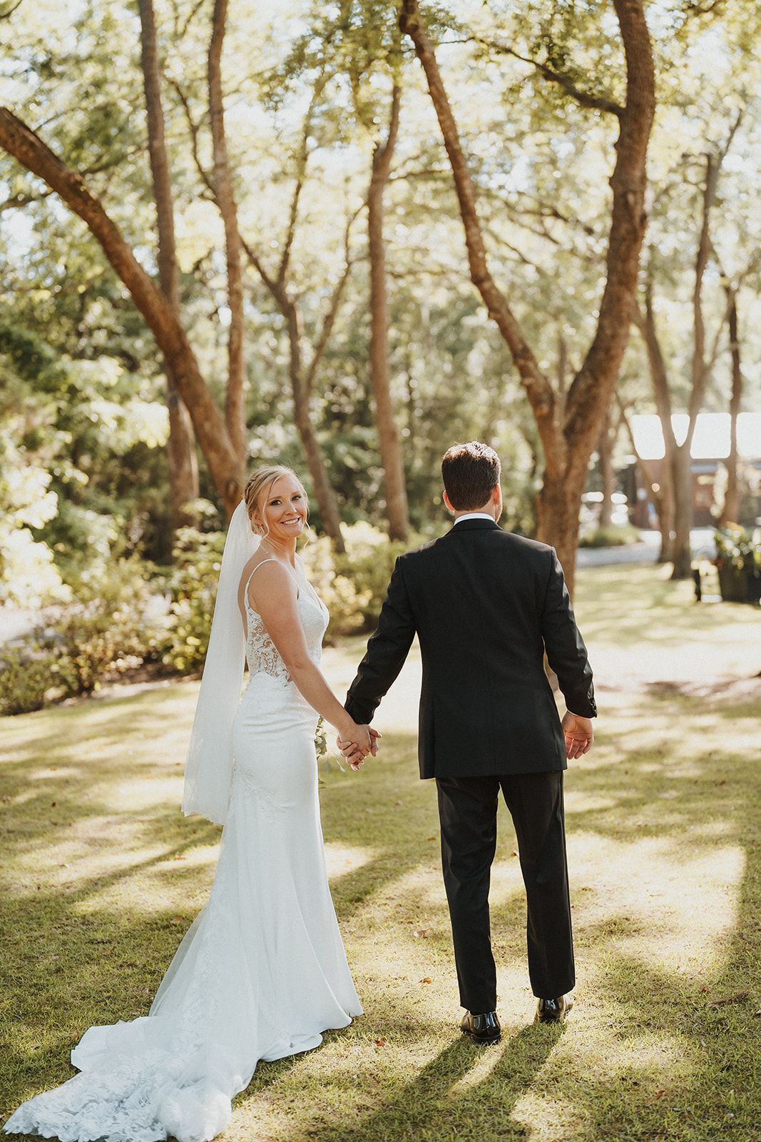 Bride and groom holding hands in a sunny wooded garden, walking away on a grassy path.