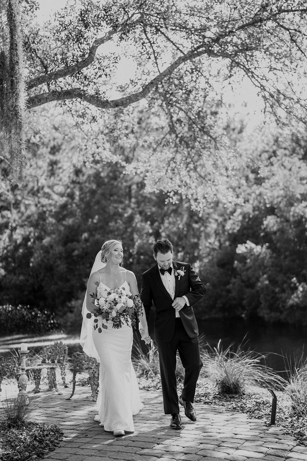 Bride and groom walking through a garden under a tree, holding hands and smiling.
