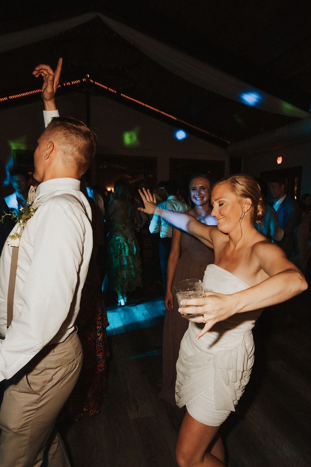 People dancing at a dimly lit party, with a woman in a white dress smiling in the foreground.