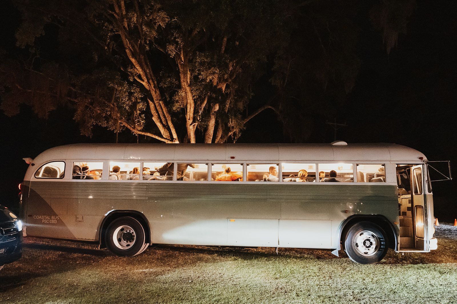 Vintage bus parked at night with warm lights glowing inside, beside a tree and grass.
