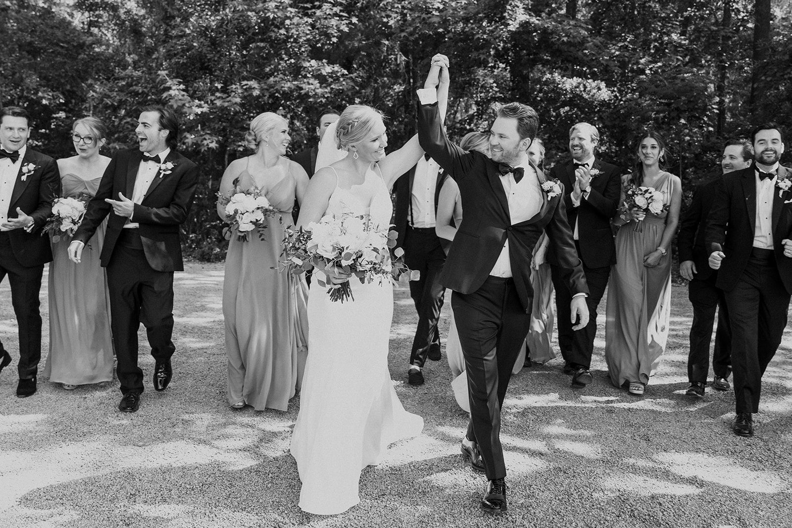 Wedding party walking down an outdoor path, with the bride and groom leading and guests clapping behind them
