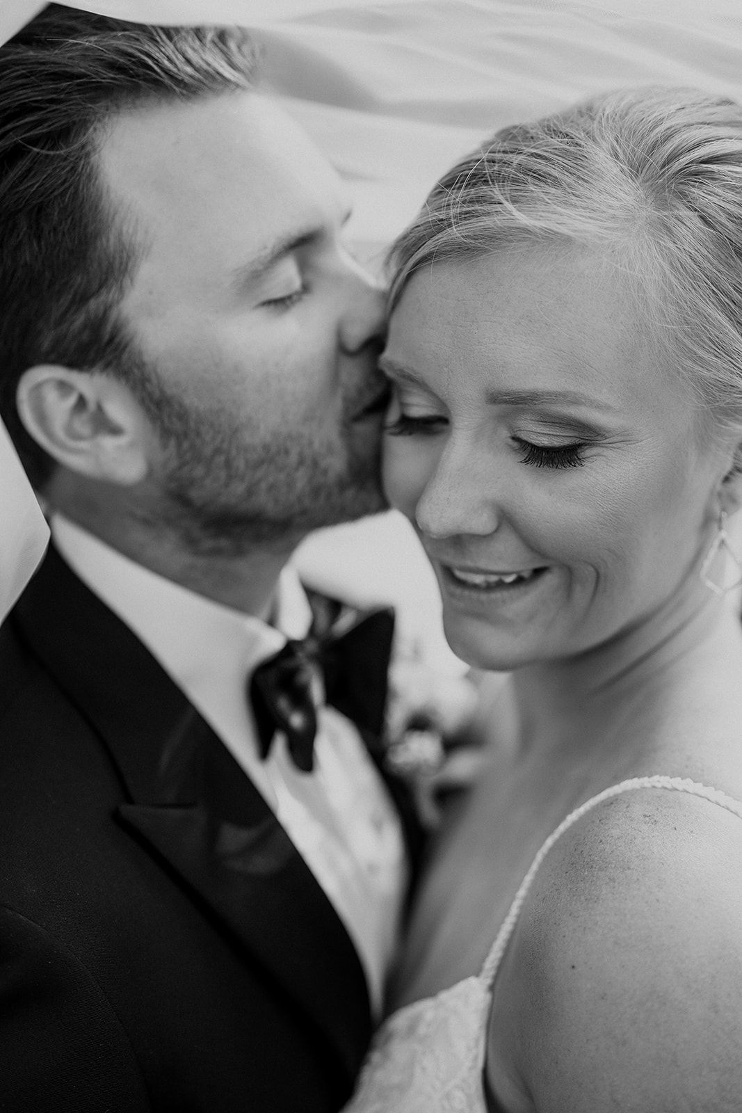 Bride and groom embracing; he kisses her cheek in a black-and-white wedding portrait.