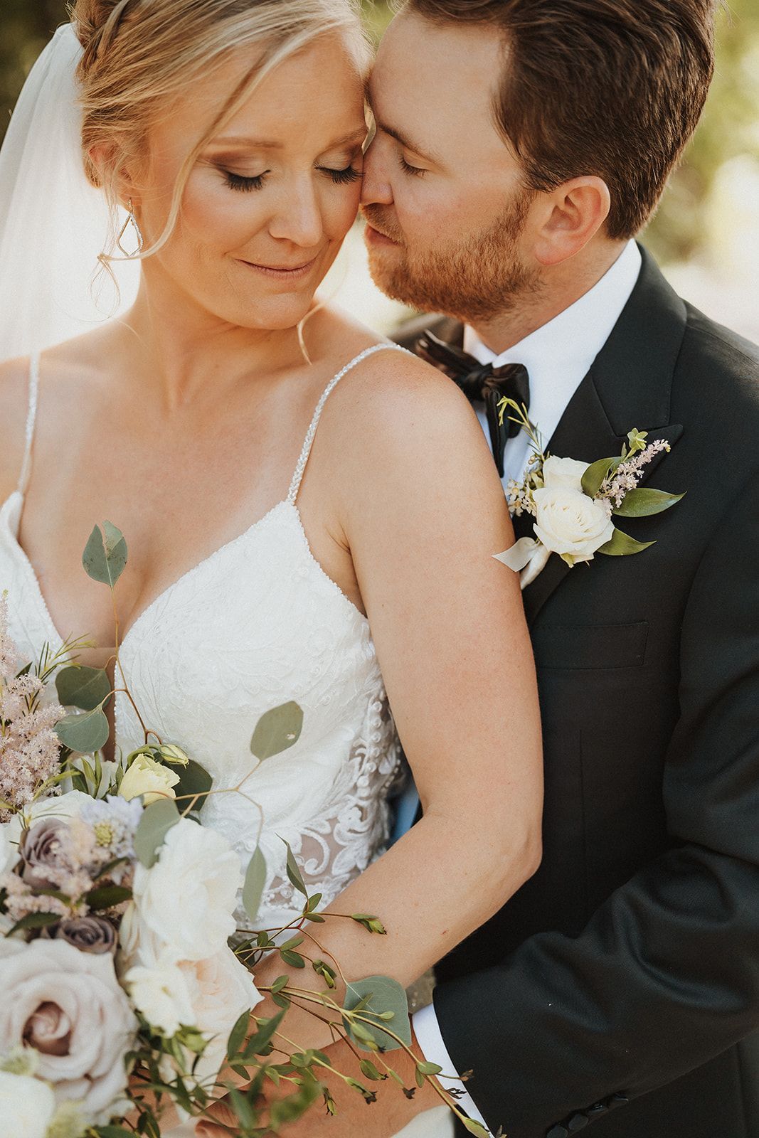 Bride and groom in wedding attire sharing a tender embrace, holding a bouquet of white flowers.