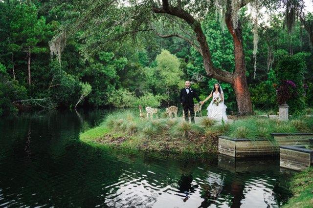 Bride and groom standing by a pond in a lush garden, surrounded by trees and greenery