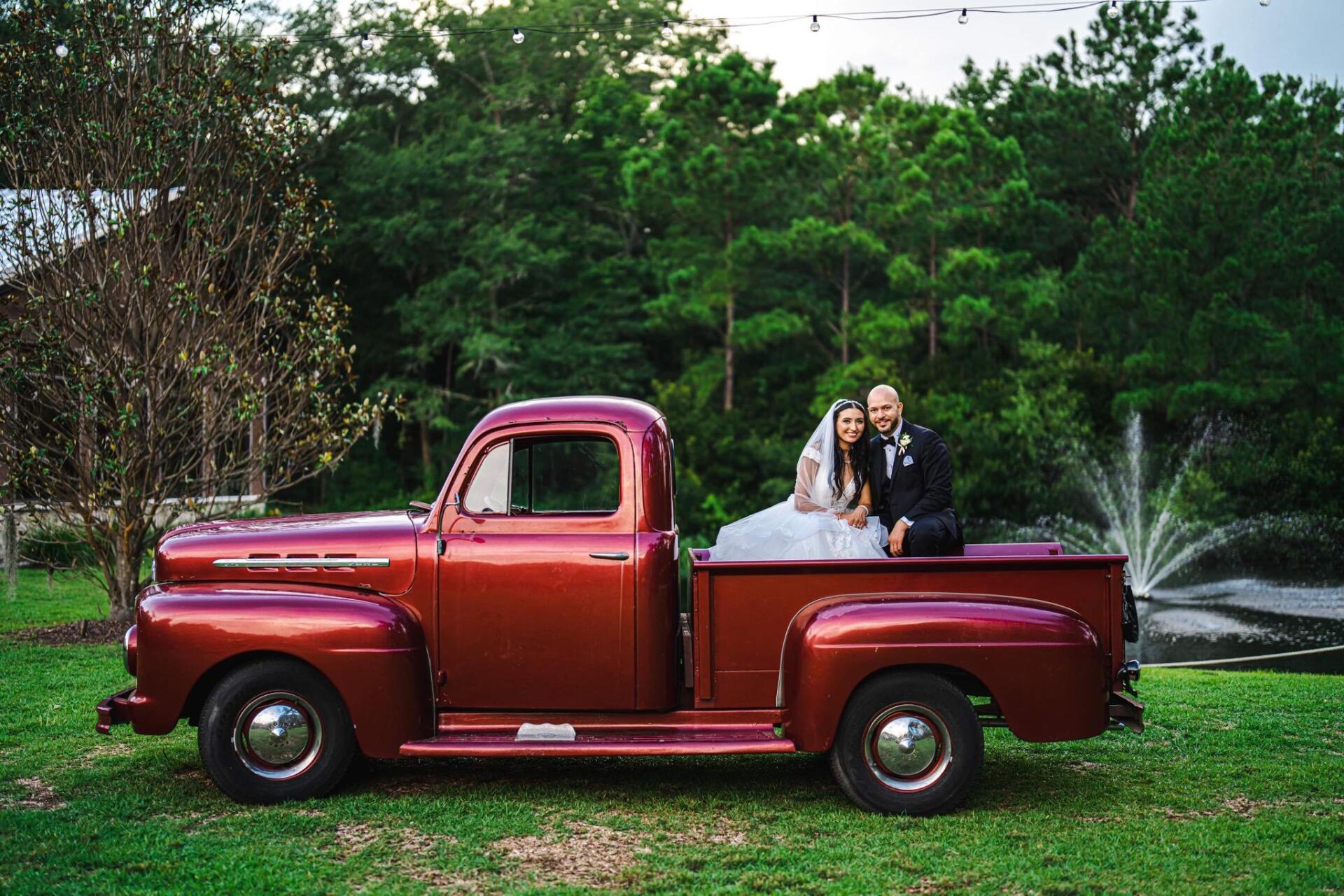 Two people in a red vintage pickup truck in a grassy park with a fountain and trees behind them.