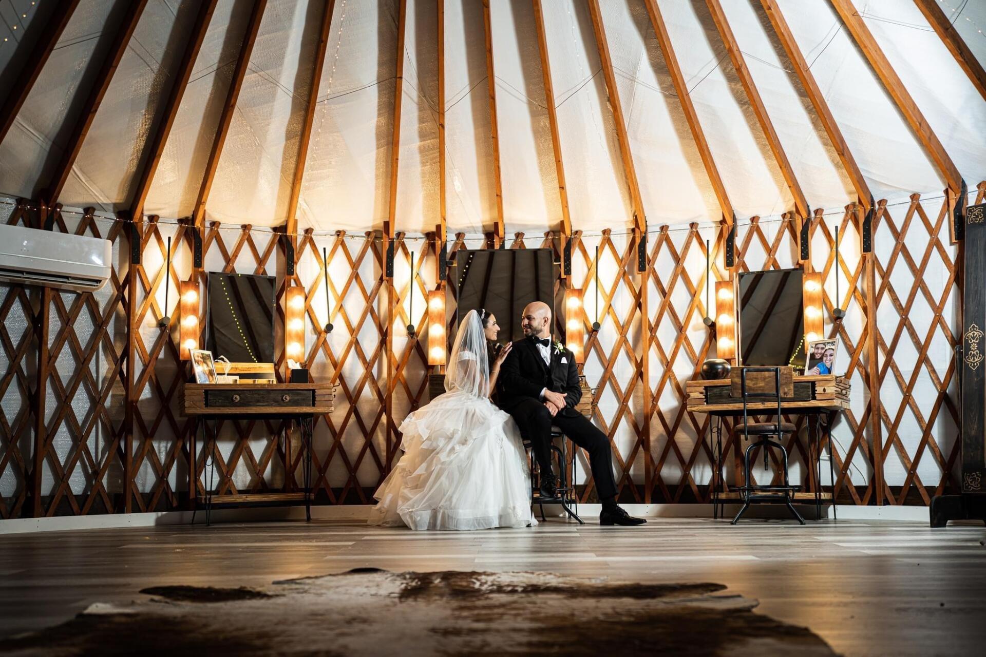 Couple seated in wedding attire inside a rustic yurt-style venue with warm geometric lighting.