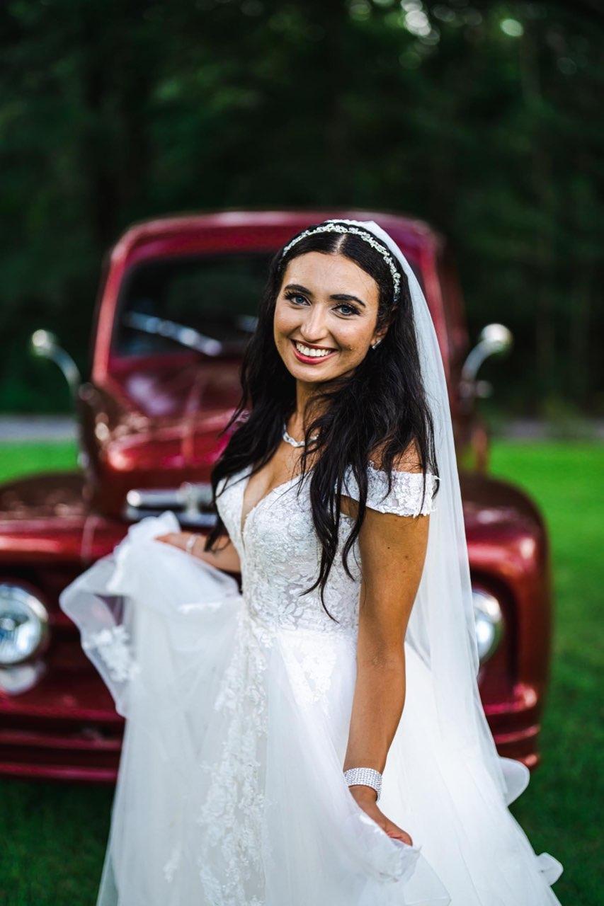 Woman in a white dress smiling in front of a vintage red car outdoors
