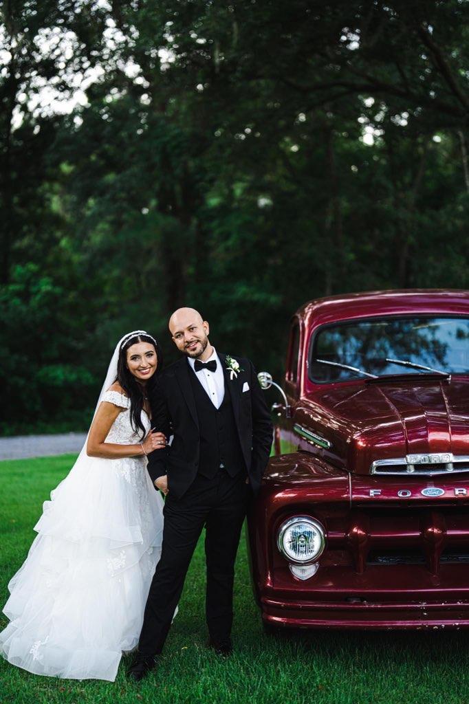 Bride and groom posing beside a burgundy classic car outdoors at dusk
