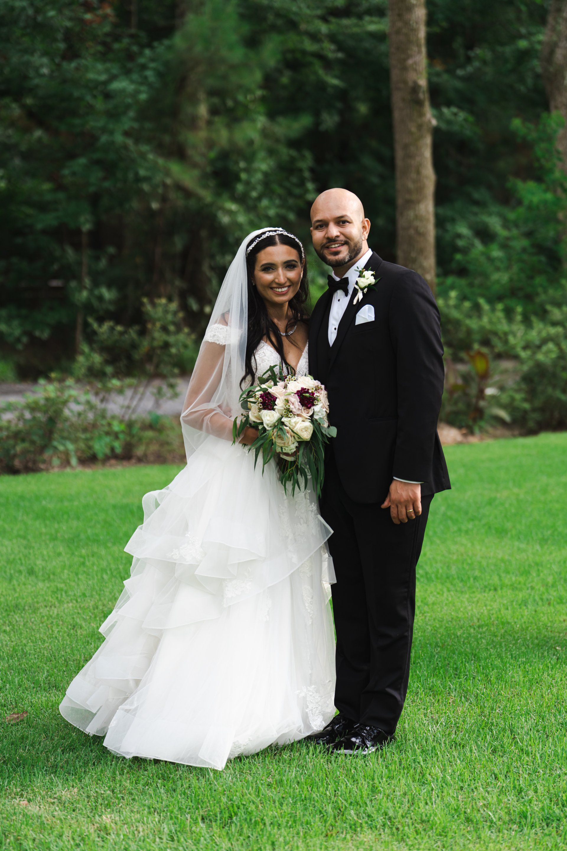 Bride and groom posing outdoors on a green lawn, bride in white gown holding bouquet, groom in black tuxedo