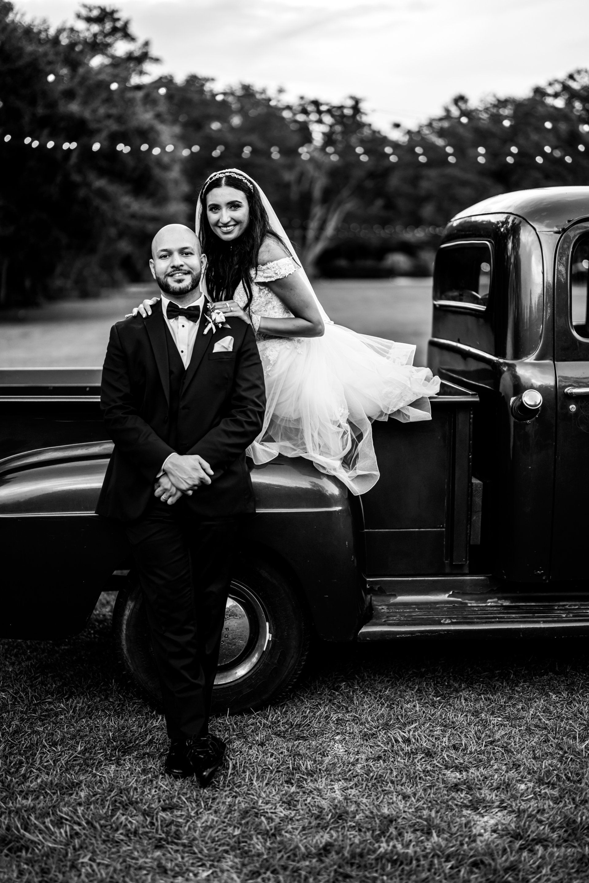 Wedding couple sitting on a vintage truck, smiling in a black-and-white outdoor portrait