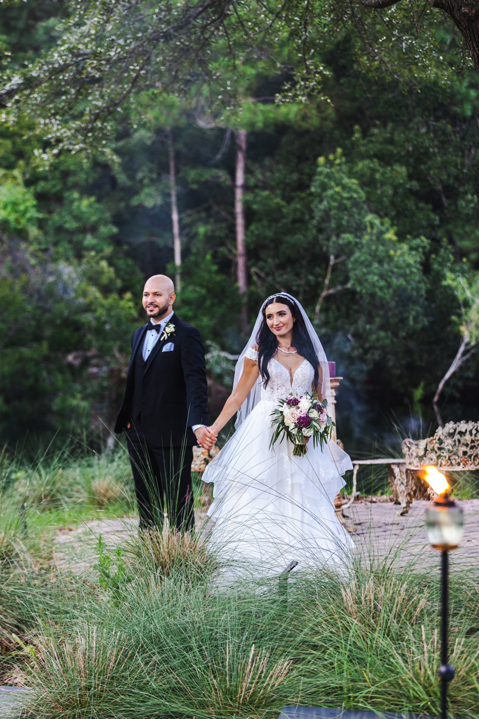 Bride and groom walking hand in hand through a grassy garden path at dusk