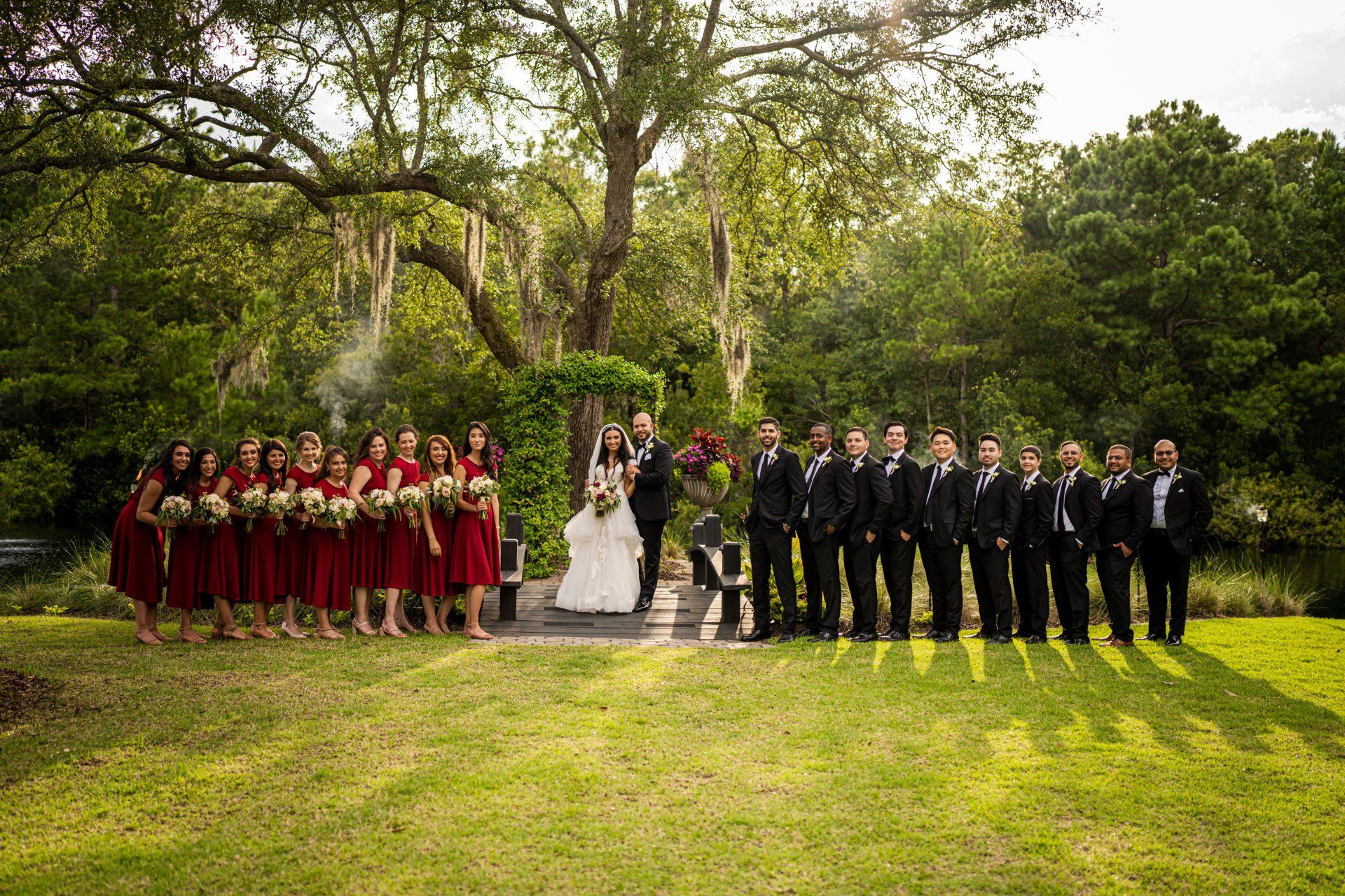 Wedding party posing outdoors on a grassy lawn, with bridesmaids in burgundy and groomsmen in dark suits.
