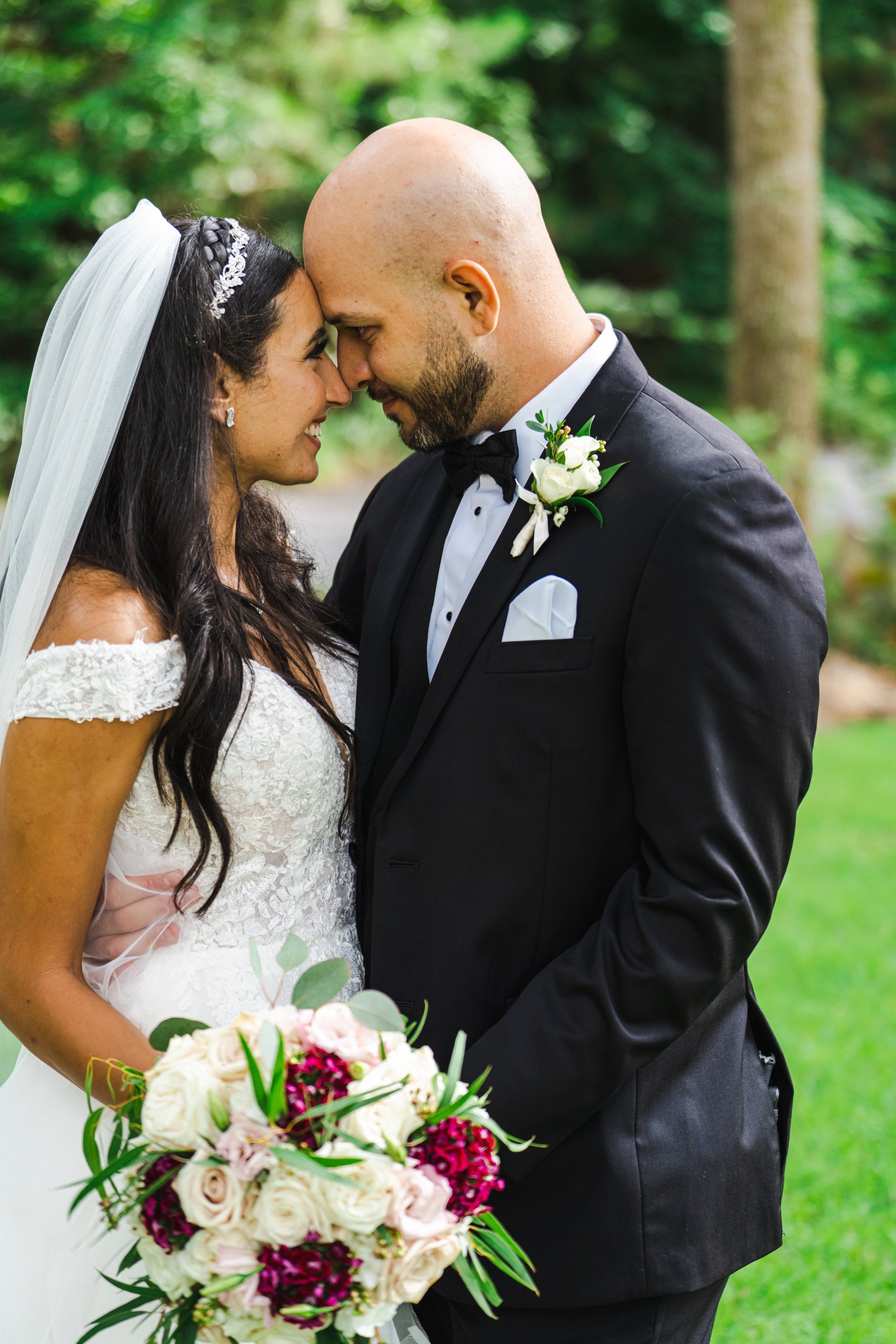 Wedding couple holding a bouquet and forehead to forehead in a green outdoor setting