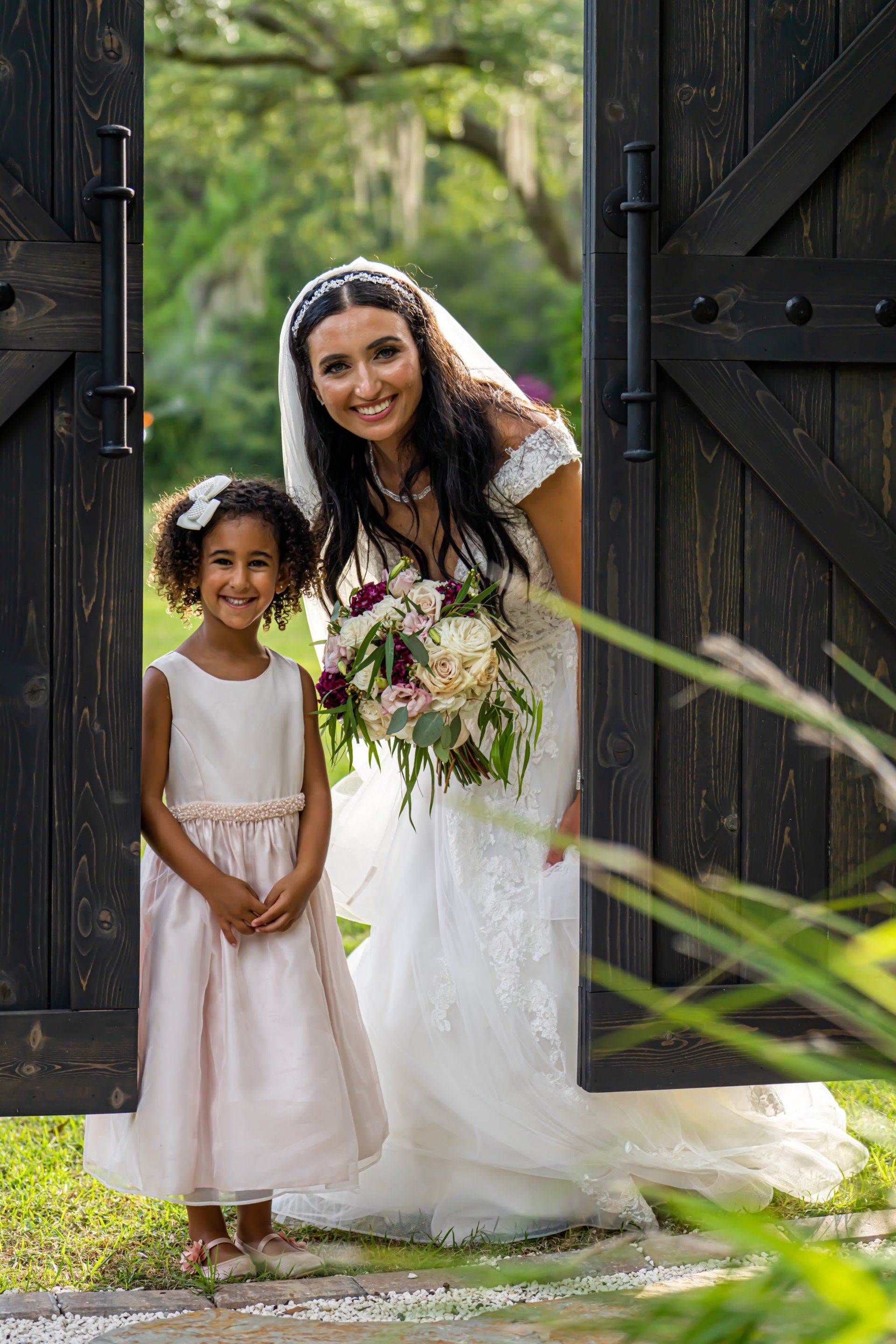 Bride and young girl standing in a doorway with a bouquet, framed by dark wooden doors and greenery.