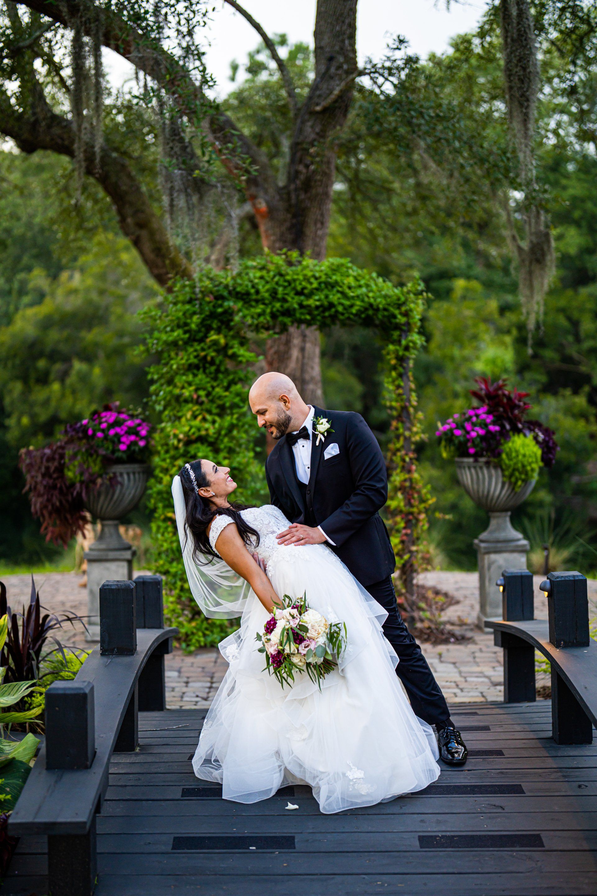 Wedding couple kissing on a wooden bridge in a garden, with purple flowers and trees nearby