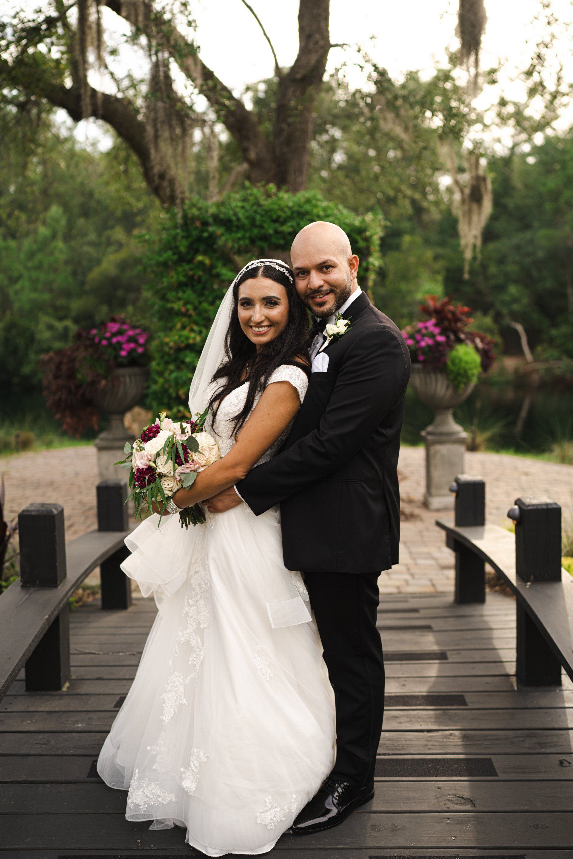 Newlywed couple posing on a wooden bridge in a garden, holding a bouquet.