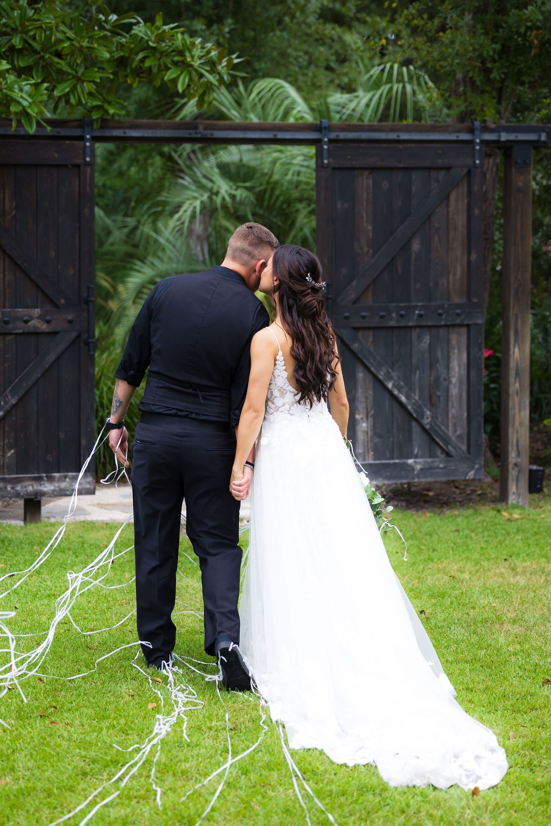 Bride and groom standing on grass before rustic black gates, sharing a kiss