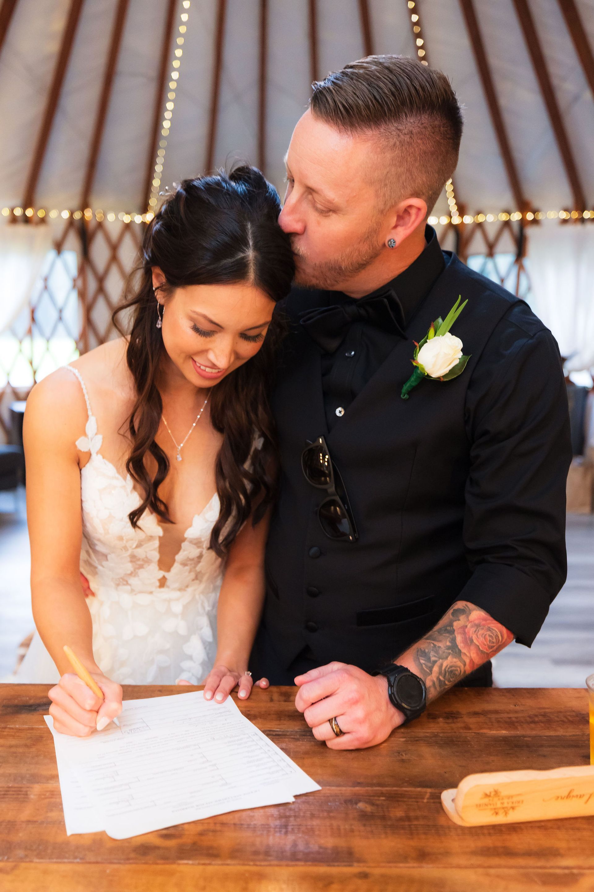 Couple signing a document at a wedding ceremony under a tent, groom leaning in affectionately.