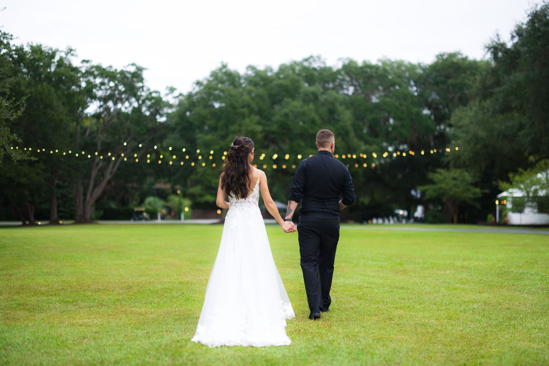 Couple in wedding attire holding hands on a grassy lawn with string lights in the background