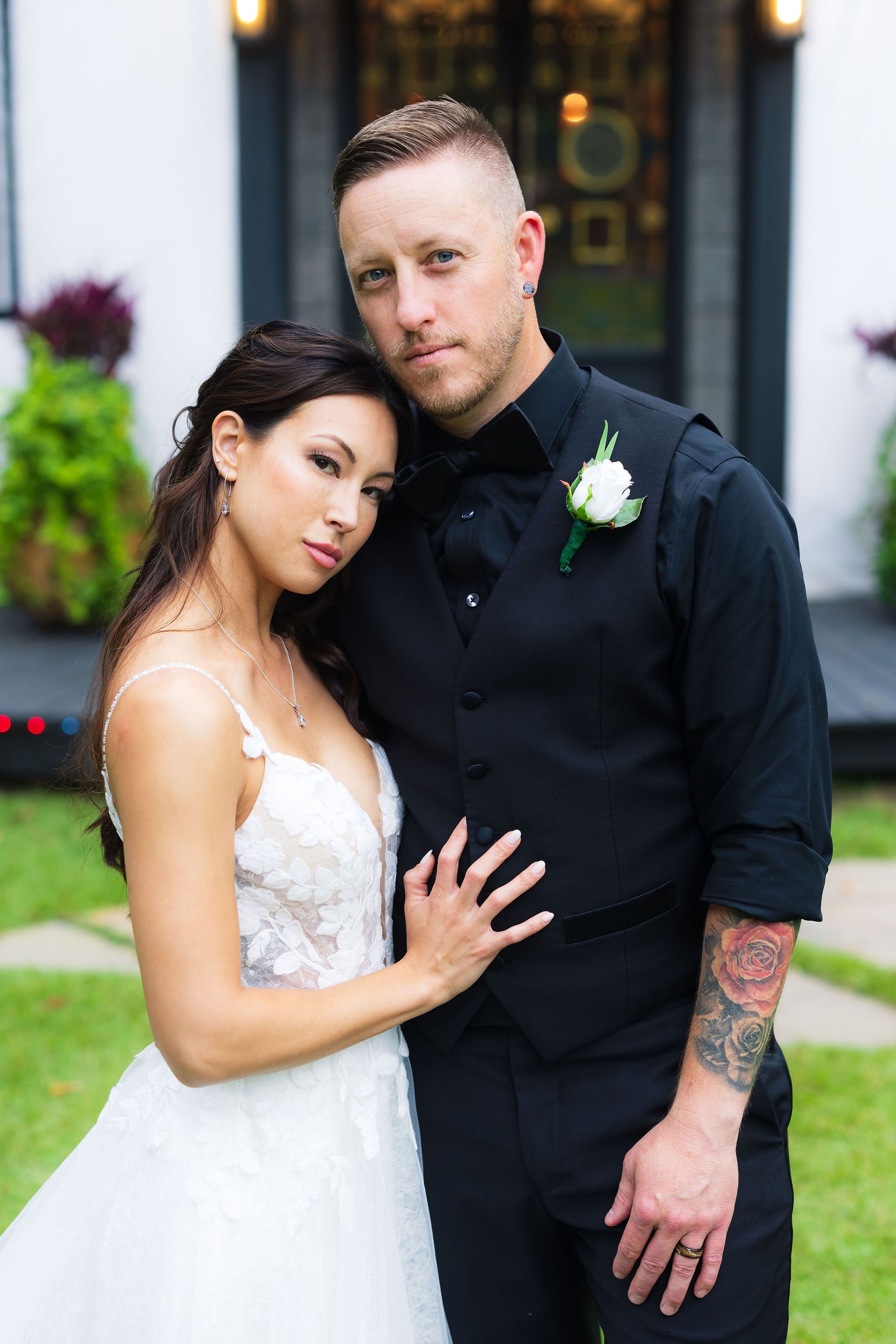 Bride in white gown and partner in black shirt posing outdoors, leaning close together.