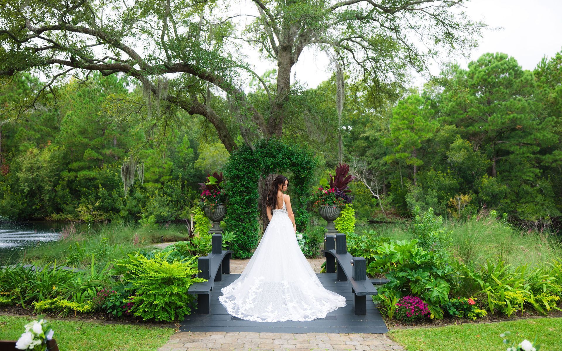 Bride in a white gown standing on a stone platform in a lush garden by the water